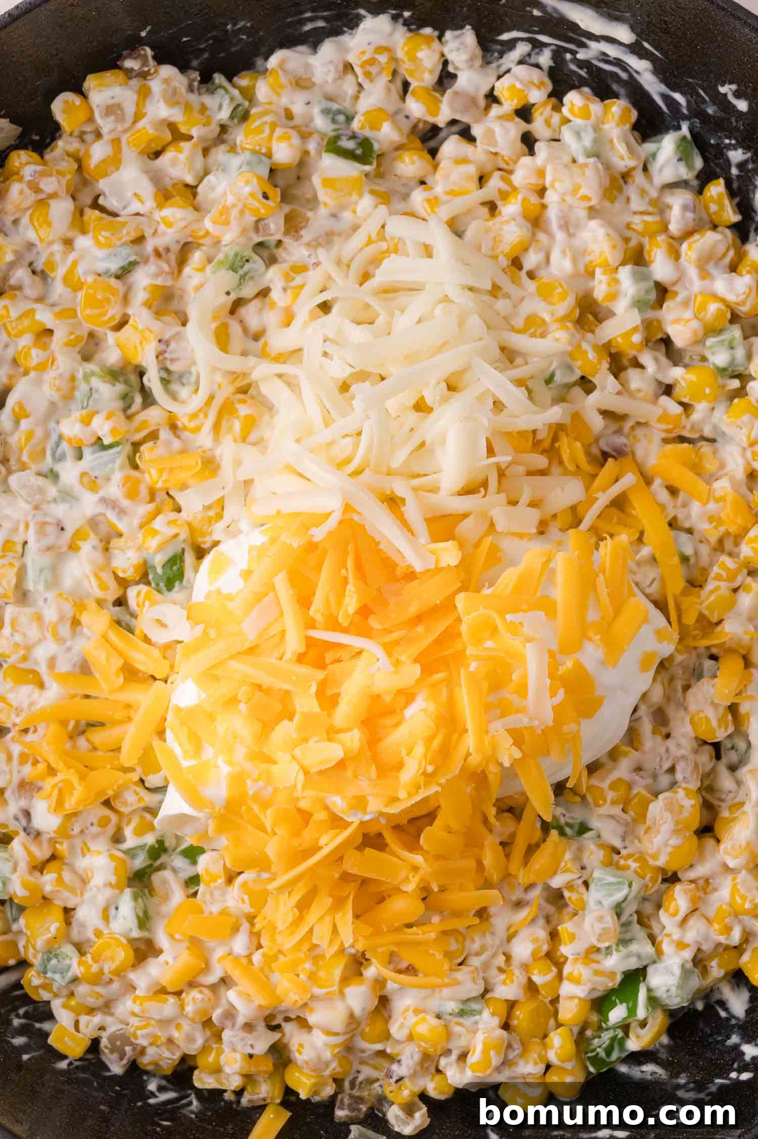 Close-up of a creamy corn mixture topped with shredded white and yellow cheese in a pan.
