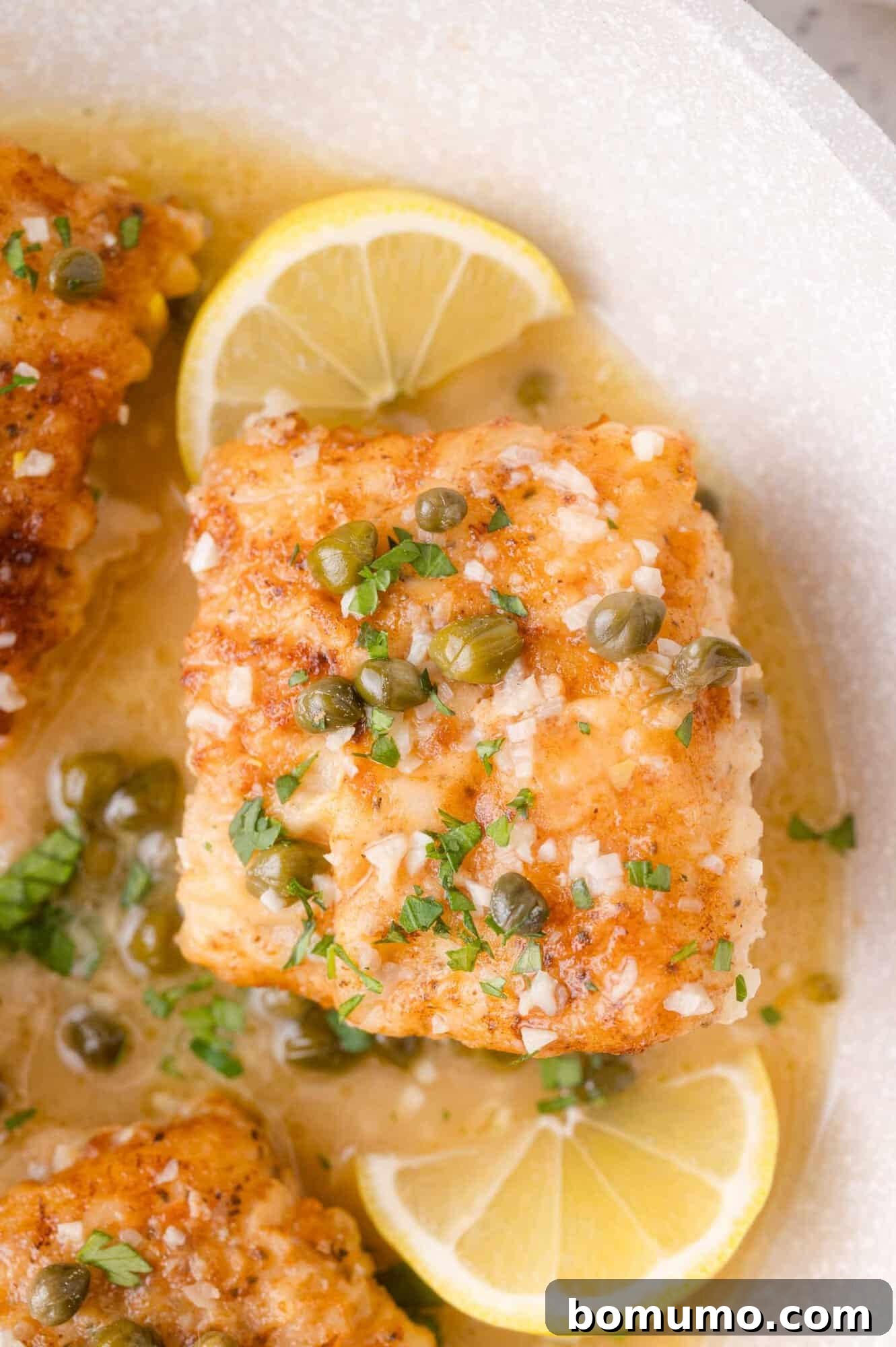Lemon-Caper Cod Piccata 2 A piece of cooked cod topped with capers and herbs, served in a light sauce with lemon wedges on a white plate.