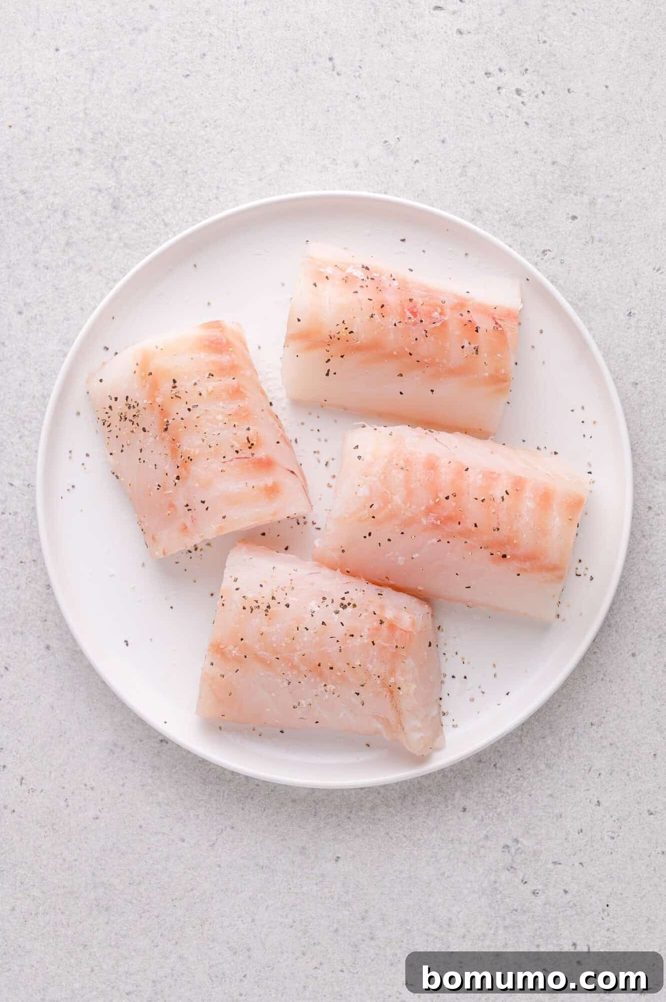 Lemon-Caper Cod Piccata 4 Raw cod fillets seasoned with black pepper on a white plate.