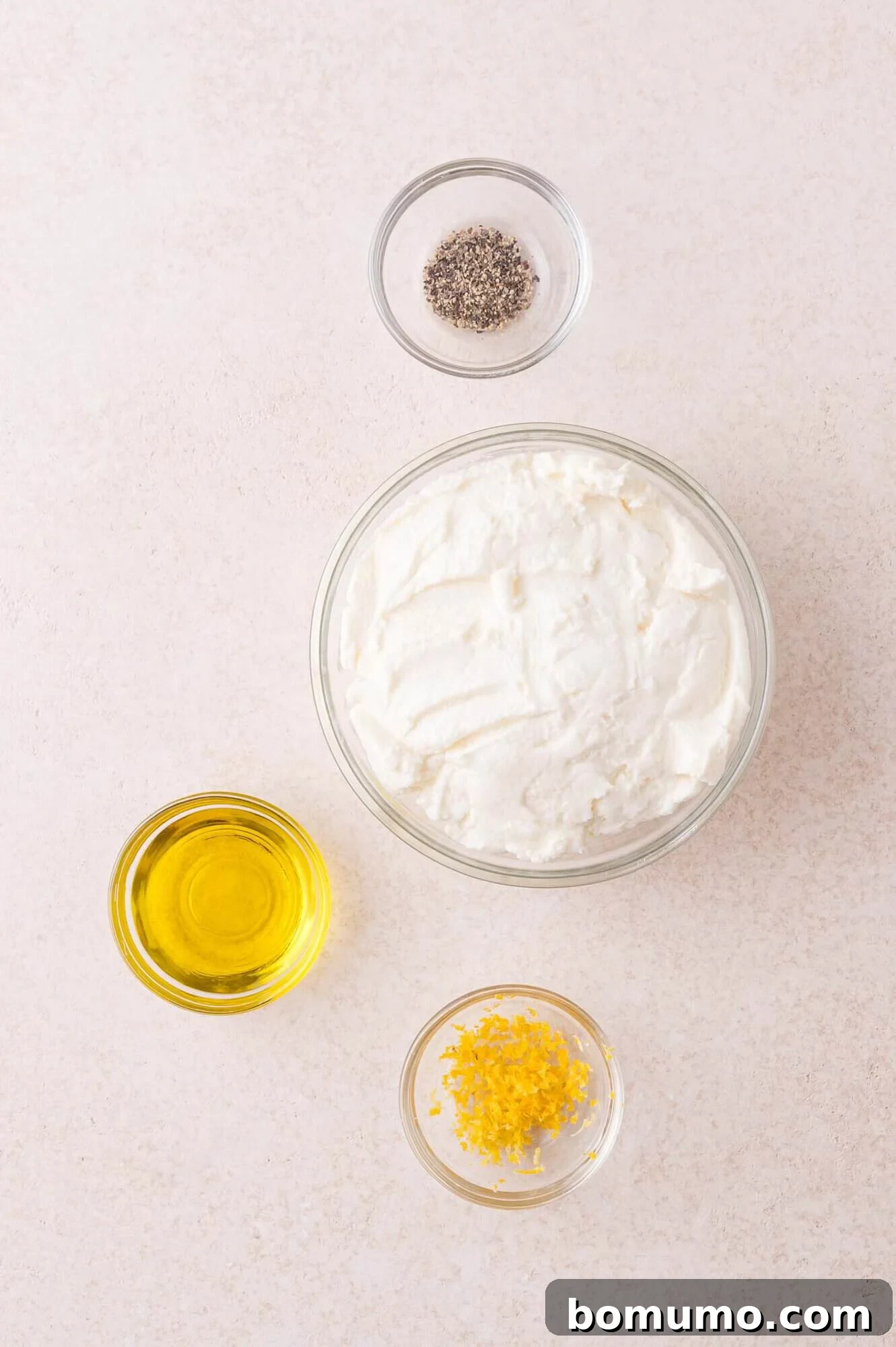 Four small bowls containing black pepper, ricotta cheese, olive oil, and lemon zest are arranged on a light-colored surface.
