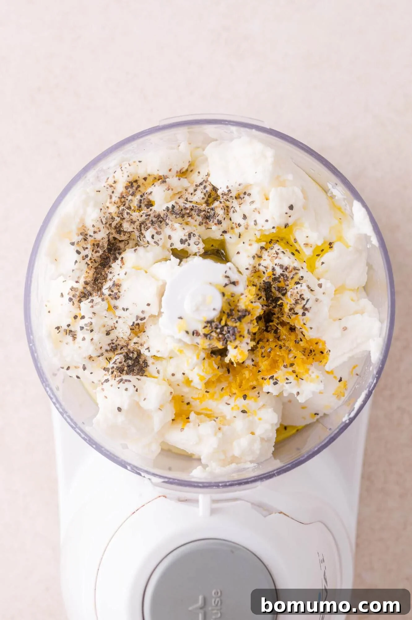 A food processor bowl filled with ricotta cheese, olive oil, lemon zest, and cracked black pepper, ready to be blended.