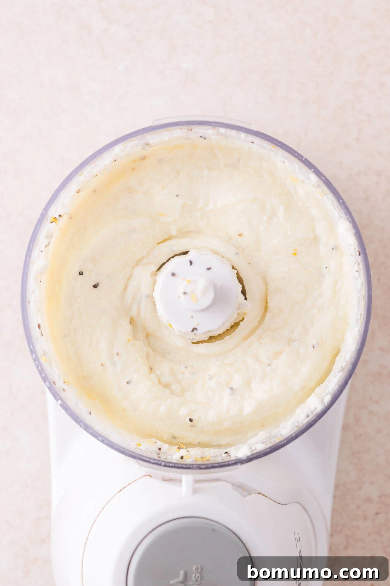 Overhead view of a food processor containing a creamy, blended mixture with visible flecks of seasoning.