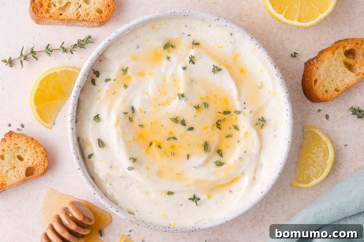 A bowl of whipped ricotta cheese topped with honey, lemon zest, and fresh thyme, surrounded by crostini, lemon slices, and a honey dipper.