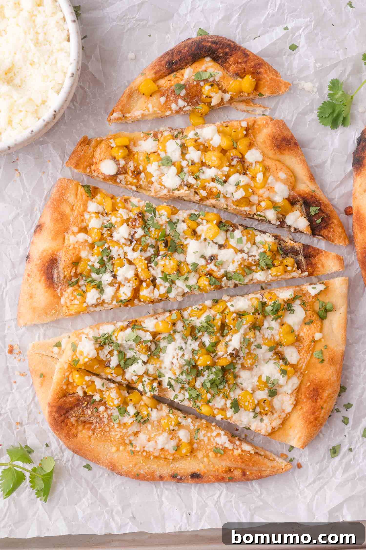 Flatbread topped with corn, cheese, herbs, and seasoning, sliced into pieces on parchment paper, with a small bowl of extra cheese nearby.