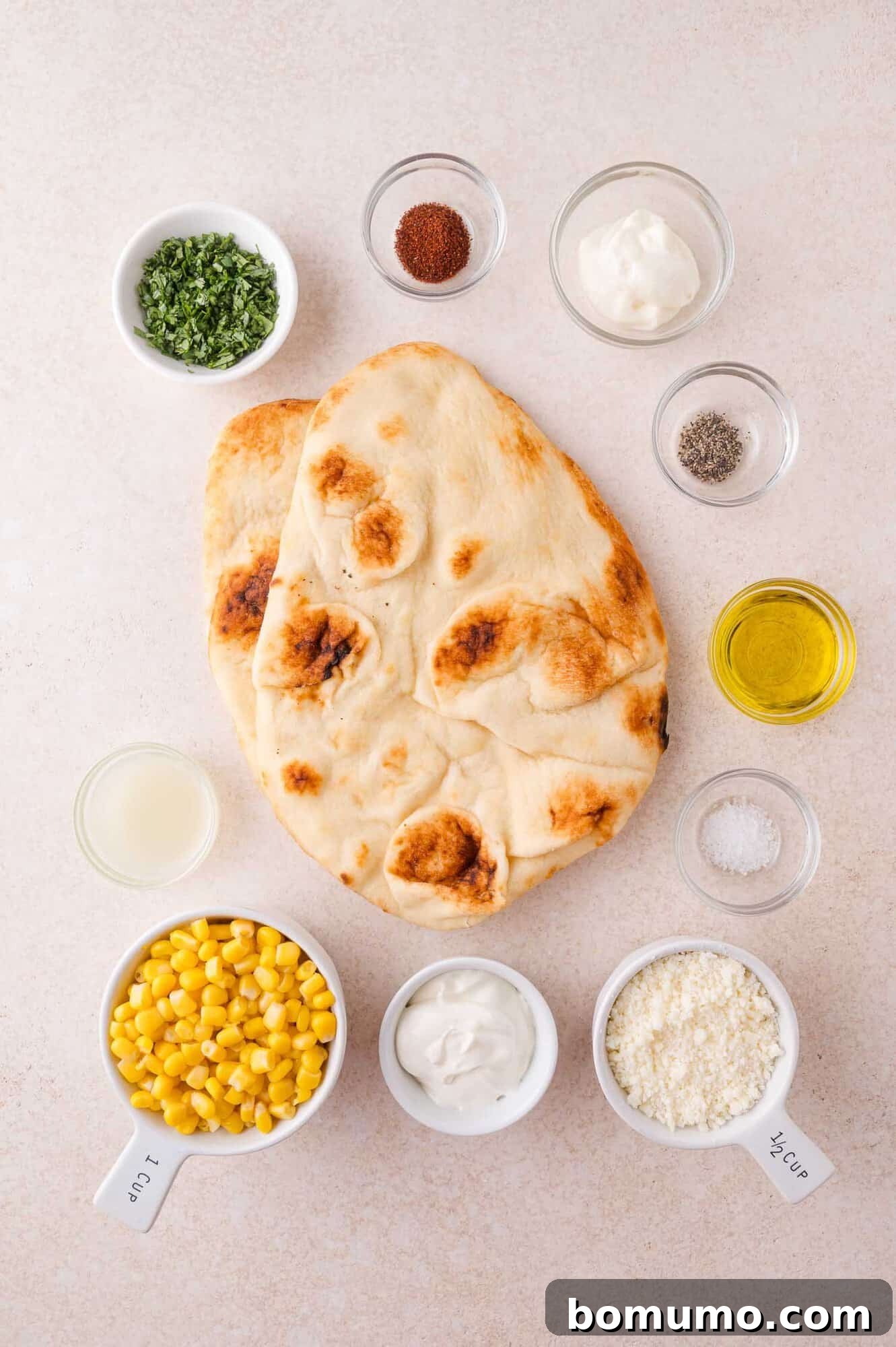 Assorted ingredients for a flatbread dish, including naan, corn, grated cheese, herbs, yogurt, mayonnaise, olive oil, spices, salt, pepper, and lemon juice in small bowls.