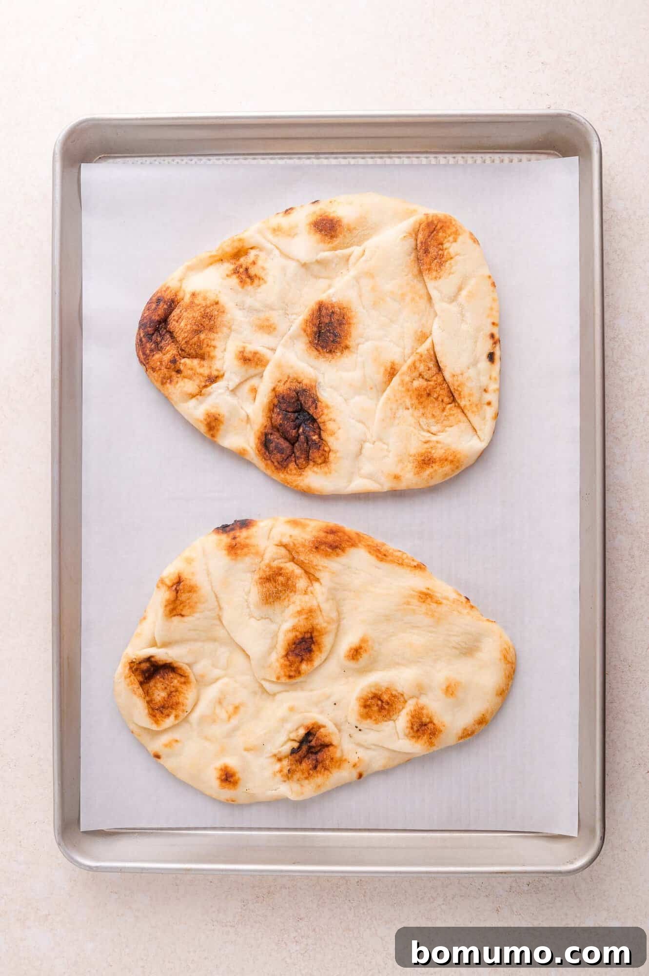 Two pieces of baked naan bread on a parchment-lined metal baking sheet.
