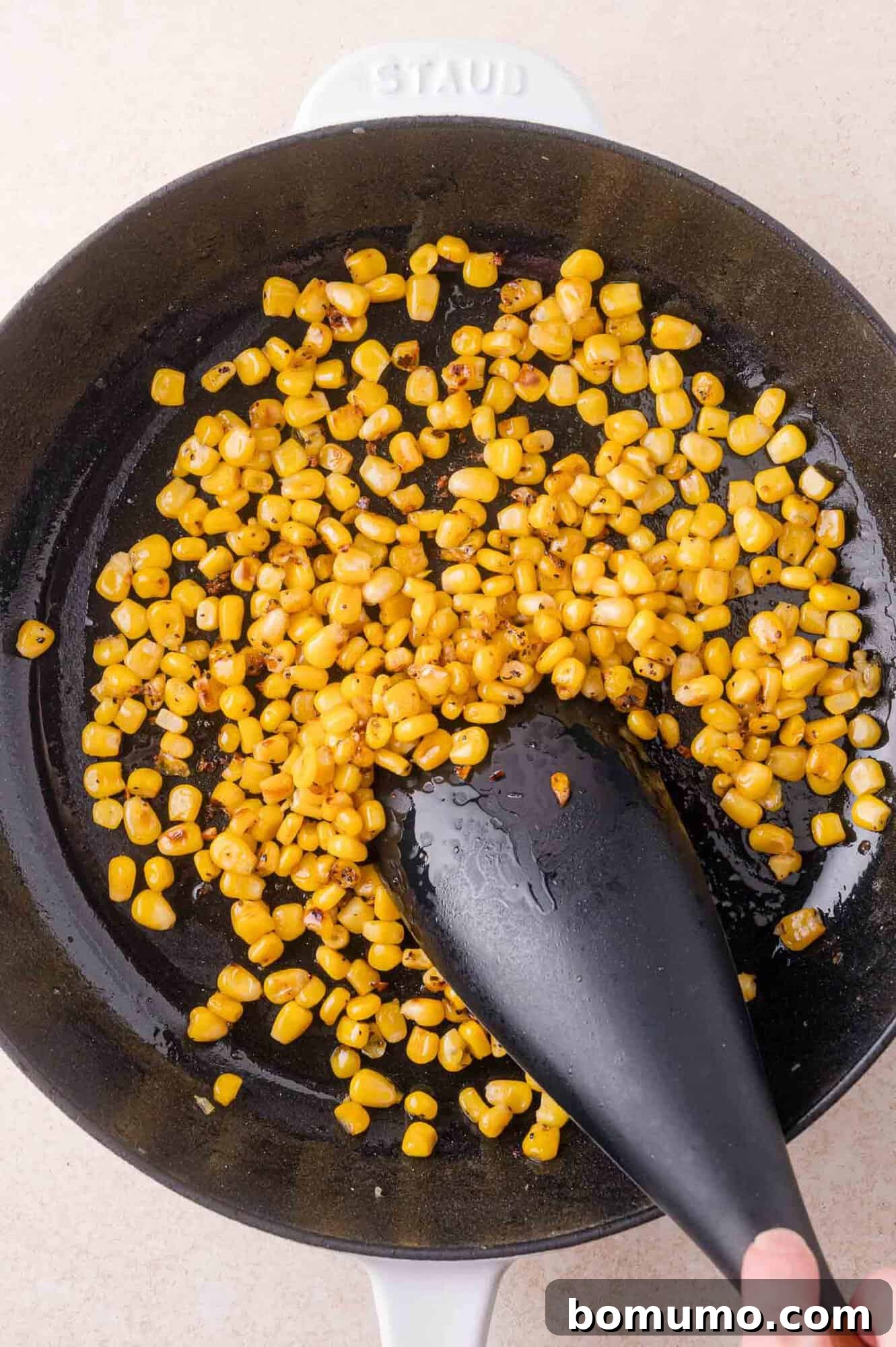 Corn kernels are being sautéed in a black pan with a black spatula; some kernels show browning from cooking.