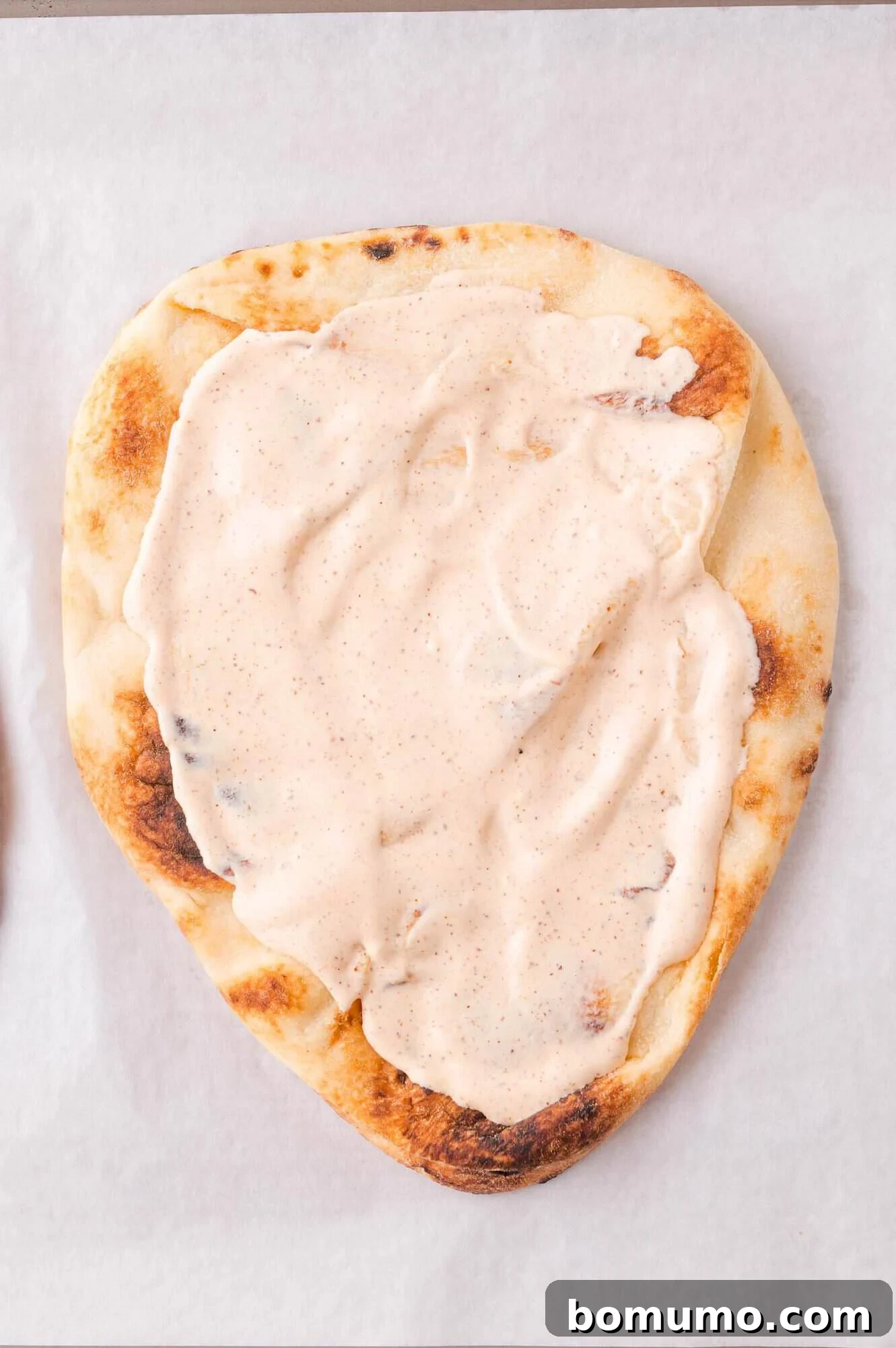 A piece of flatbread topped with a thick, creamy white sauce spread evenly over the surface, placed on white parchment paper.