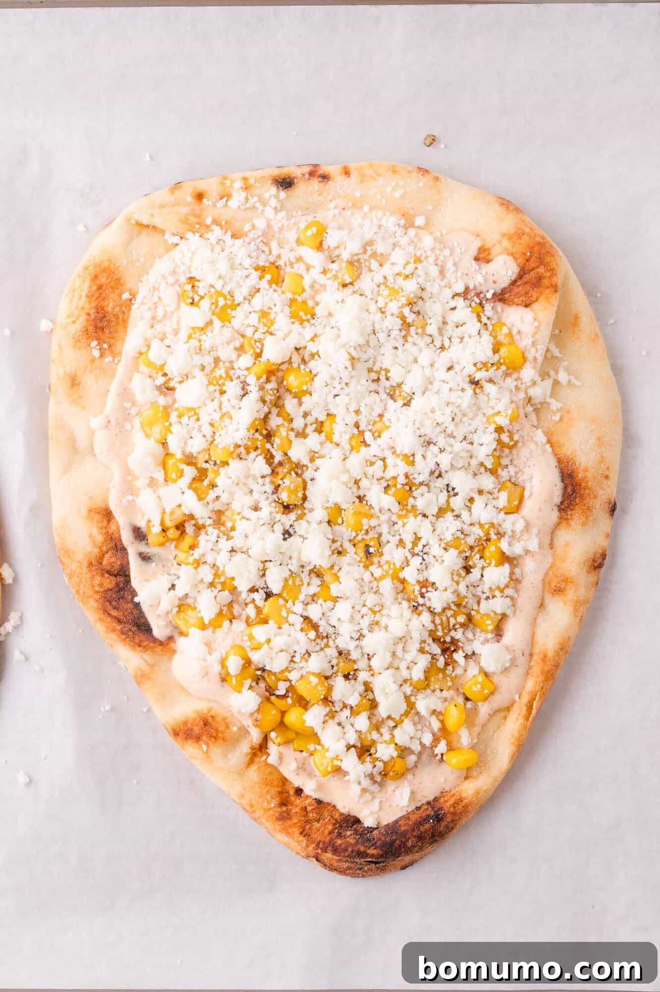 A piece of flatbread topped with a creamy sauce, corn kernels, and crumbled white cheese on a sheet of parchment paper.