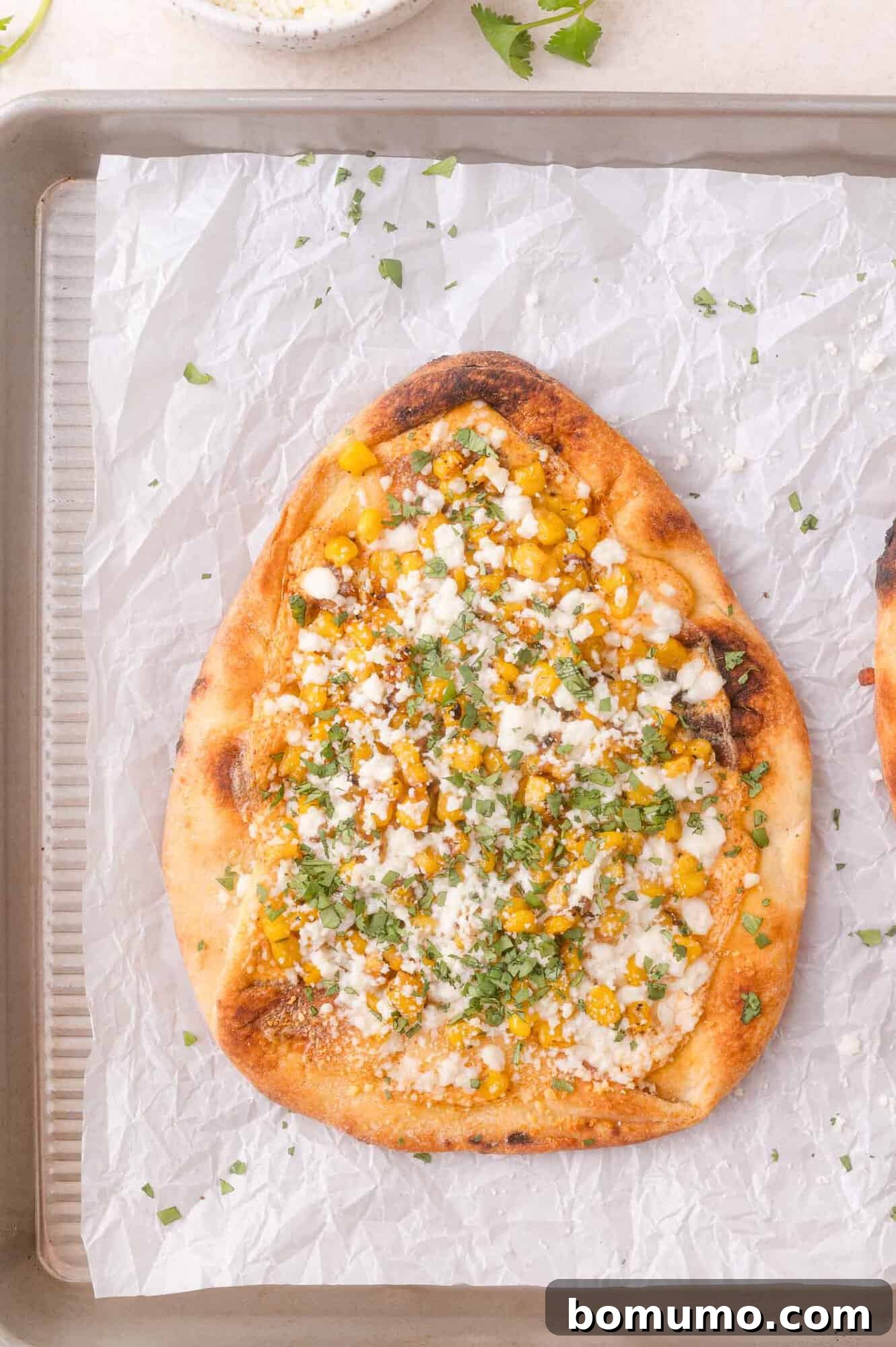 Elote flatbread topped with corn, cotija cheese, chopped cilantro, and a light sauce, served on parchment-lined baking sheet.