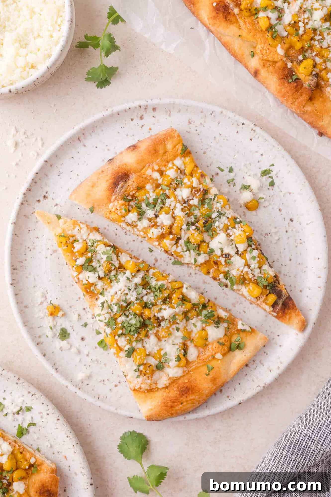 Two slices of elote flatbread topped with corn, crumbled cheese, and chopped herbs are served on a speckled white plate.