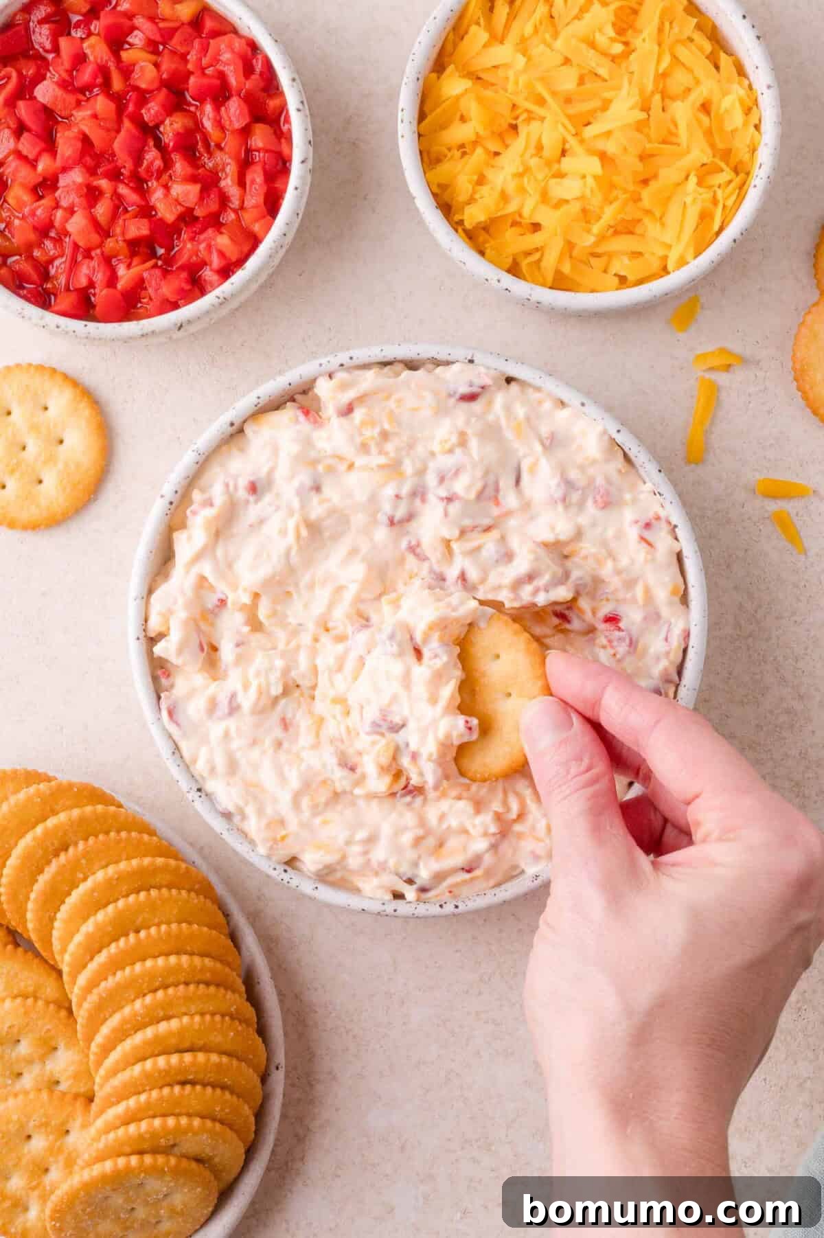 A hand dips a round cracker into a bowl of creamy pimento cheese, surrounded by bowls of shredded cheese, diced pimentos, and more crackers.