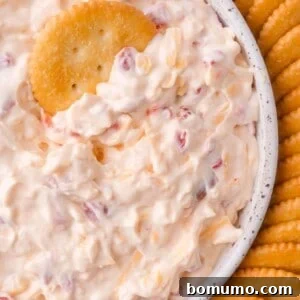 A close-up of a bowl filled with creamy pimento cheese dip containing red pimento pieces, surrounded by round crackers, with one cracker dipped in the mixture.