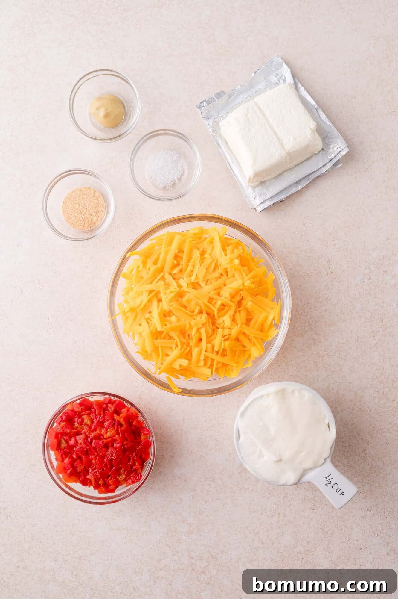 Ingredients for a cheese dip recipe arranged on a countertop, including shredded cheddar, cream cheese, diced pimentos, mayonnaise, mustard, salt, and onion powder.