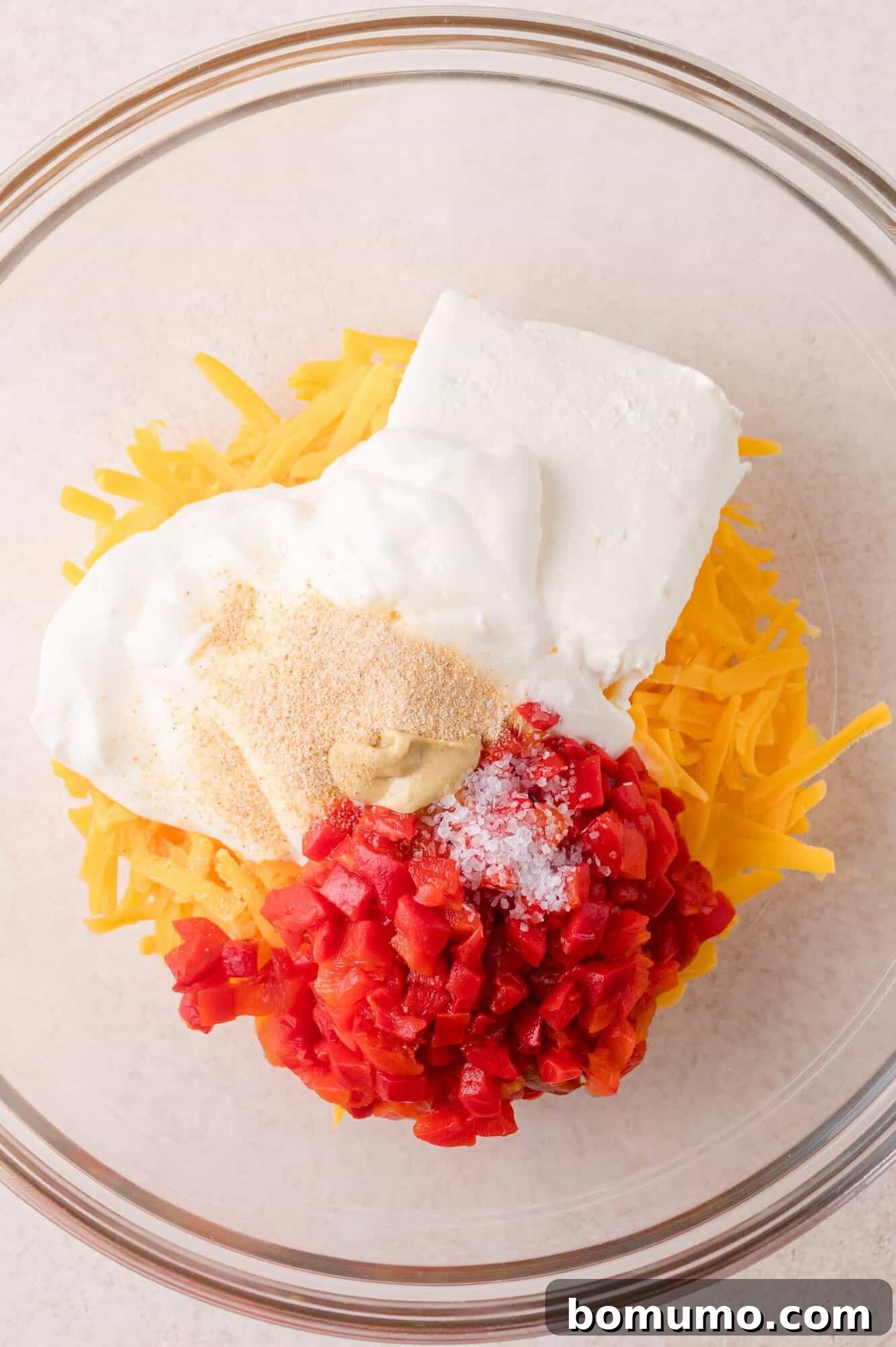 A glass bowl containing shredded cheddar cheese, cream cheese, diced pimentos, mayonnaise, mustard, garlic powder, and salt.