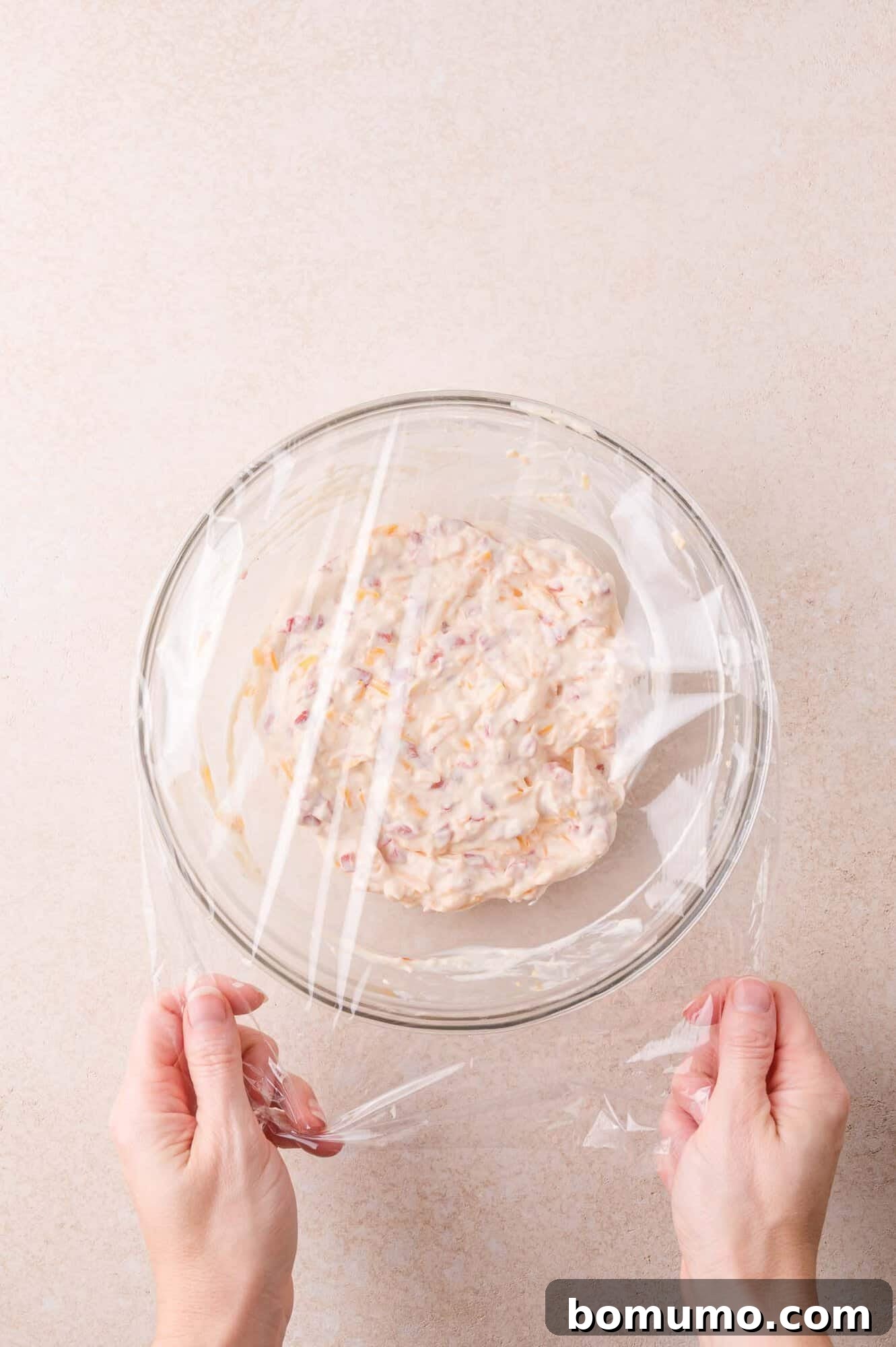 Hands covering a glass bowl filled with a pimento cheese with plastic wrap.