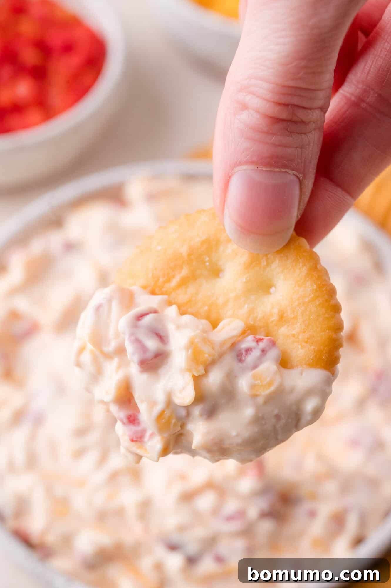 A hand holds a round cracker dipped in a creamy pimento cheese with visible bits of red and yellow ingredients.