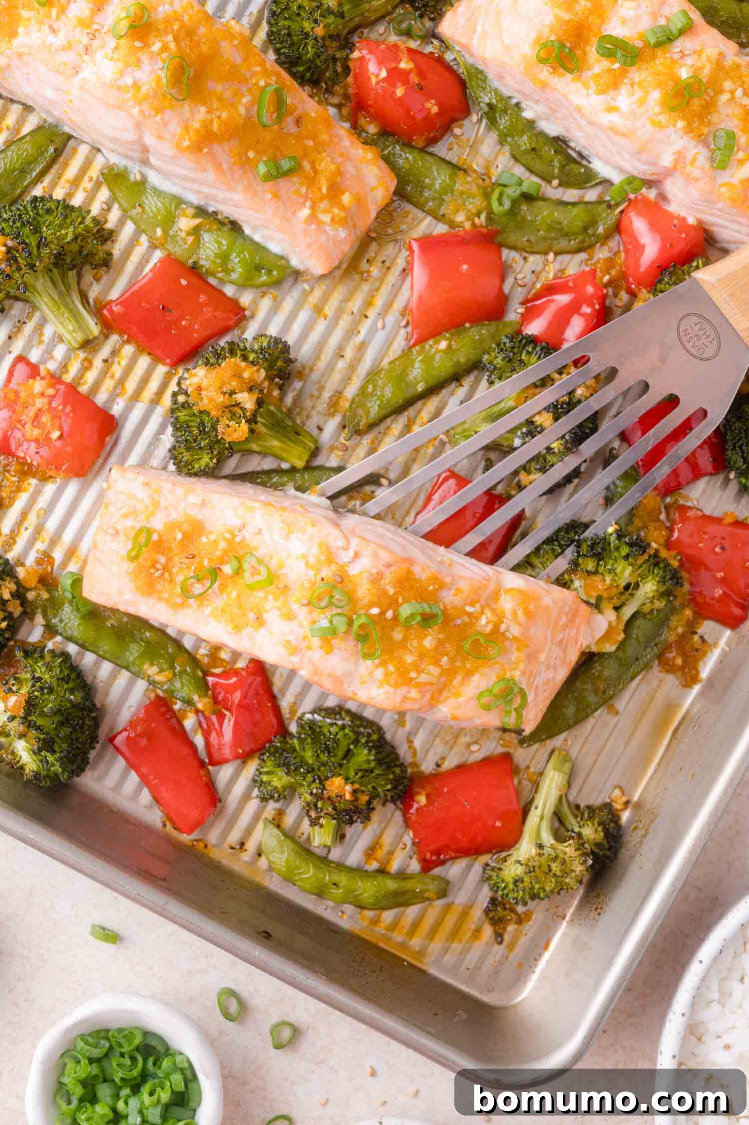 A metal spatula lifts a baked salmon fillet from a sheet pan with broccoli, red bell peppers, and snap peas.