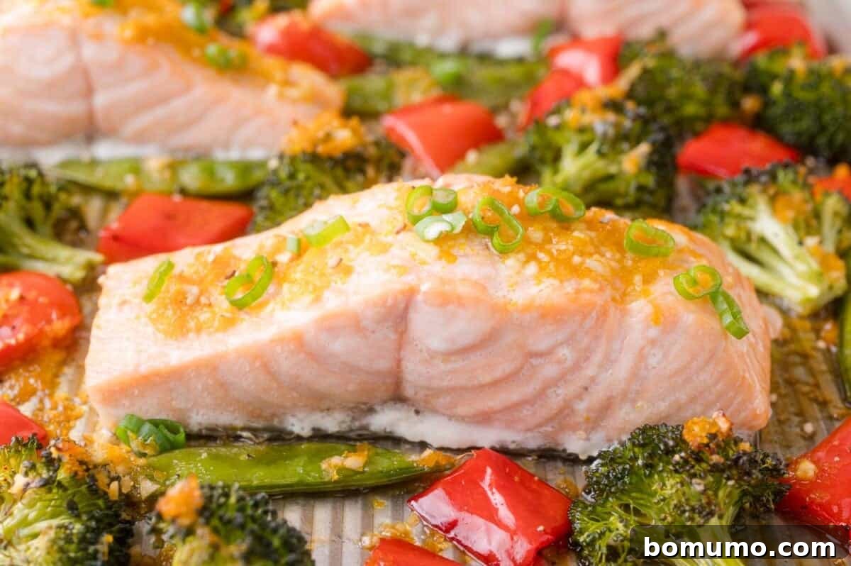 Baked salmon fillet topped with chopped green onions, surrounded by broccoli florets, red bell peppers, and snap peas on a sheet pan.