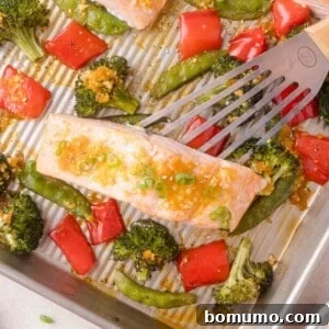A metal spatula lifts a cooked salmon fillet garnished with green onions from a baking sheet with roasted broccoli, red bell peppers, and snap peas.