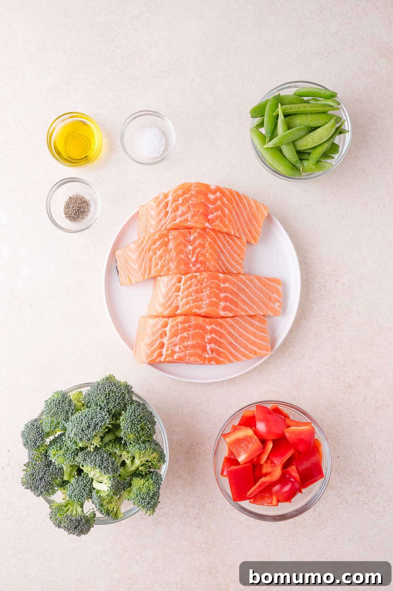 A plate of raw salmon fillets on a white surface, surrounded by bowls of broccoli, red bell pepper, snap peas, olive oil, salt, and pepper.