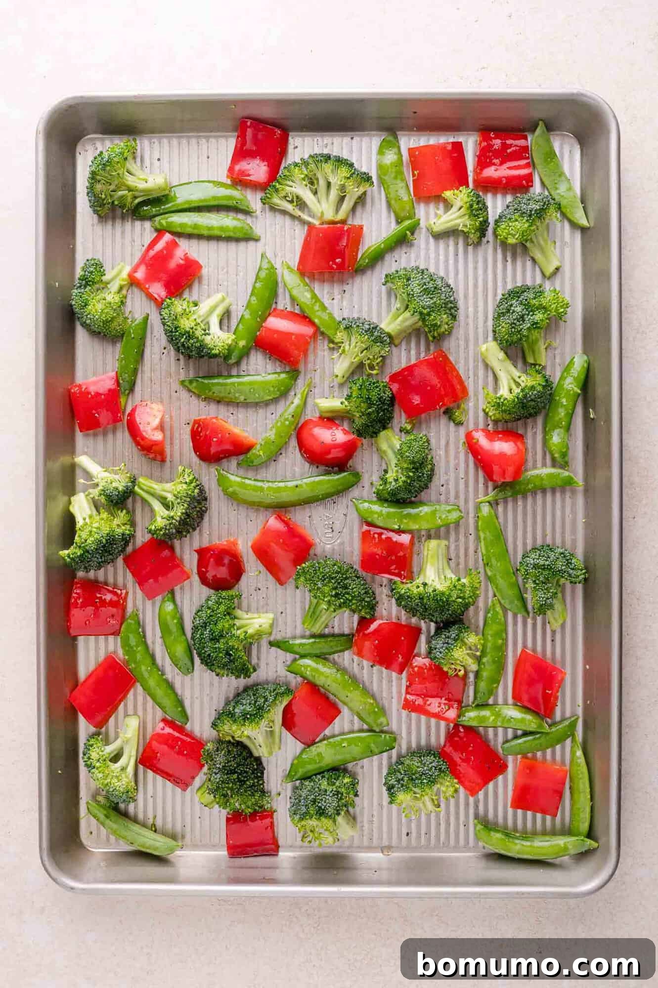 A baking sheet with raw broccoli florets, red bell pepper pieces, and snap peas spread evenly on a light-colored surface.