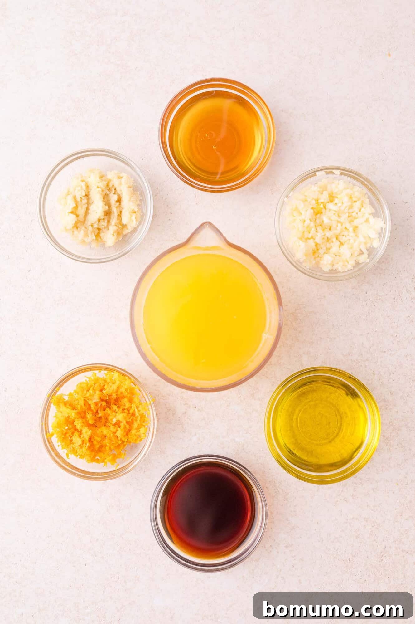 Seven small bowls containing various ingredients, including orange juice, olive oil, soy sauce, honey, grated ginger, grated garlic, and orange zest, arranged in a circle on a light surface.