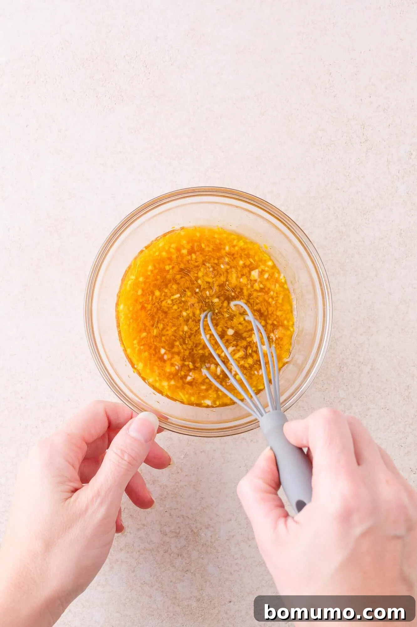A person whisking a yellow-brown liquid mixture in a clear glass bowl on a light-colored surface.