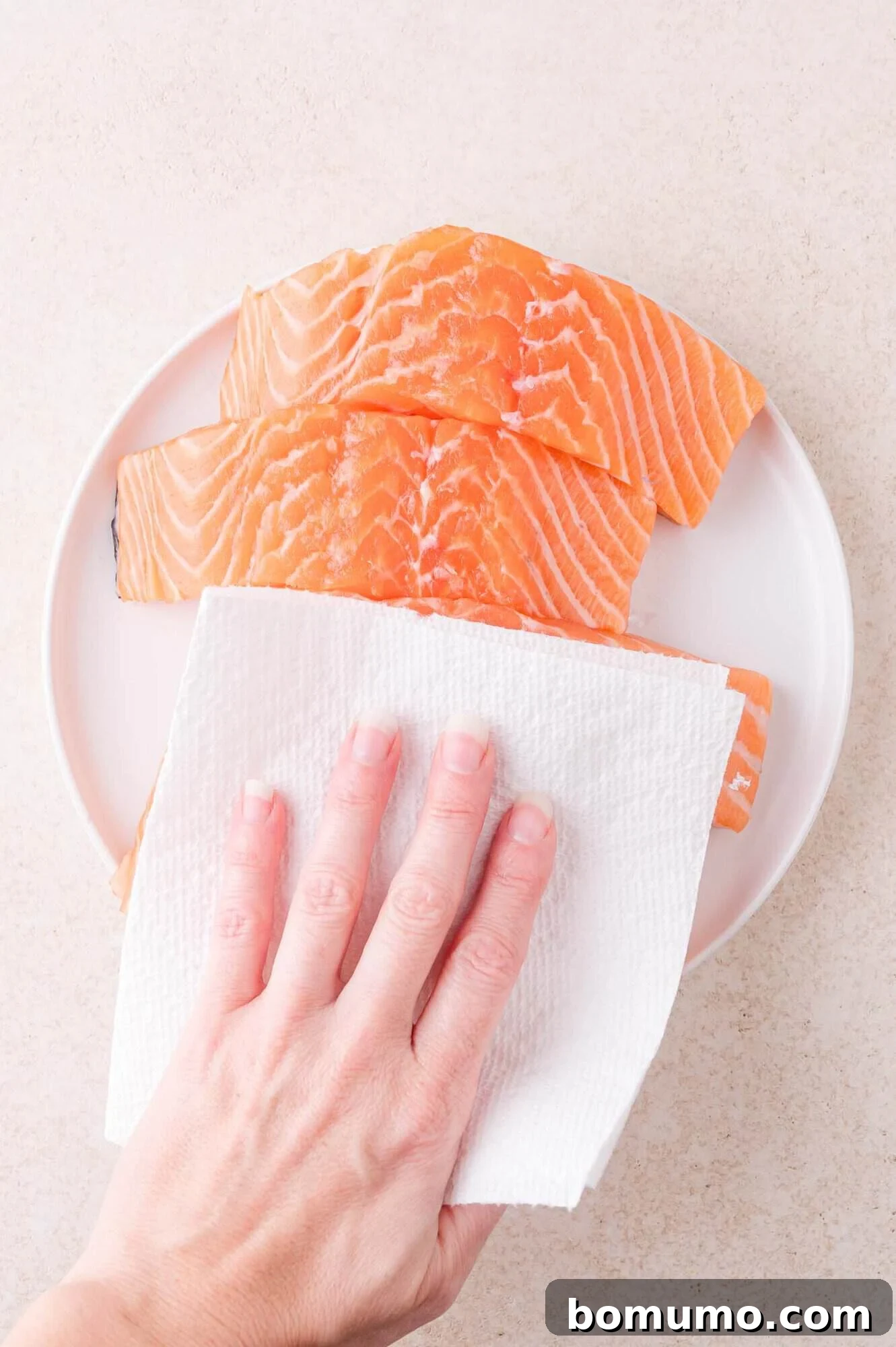 A hand uses a paper towel to pat dry raw salmon fillets on a white plate.