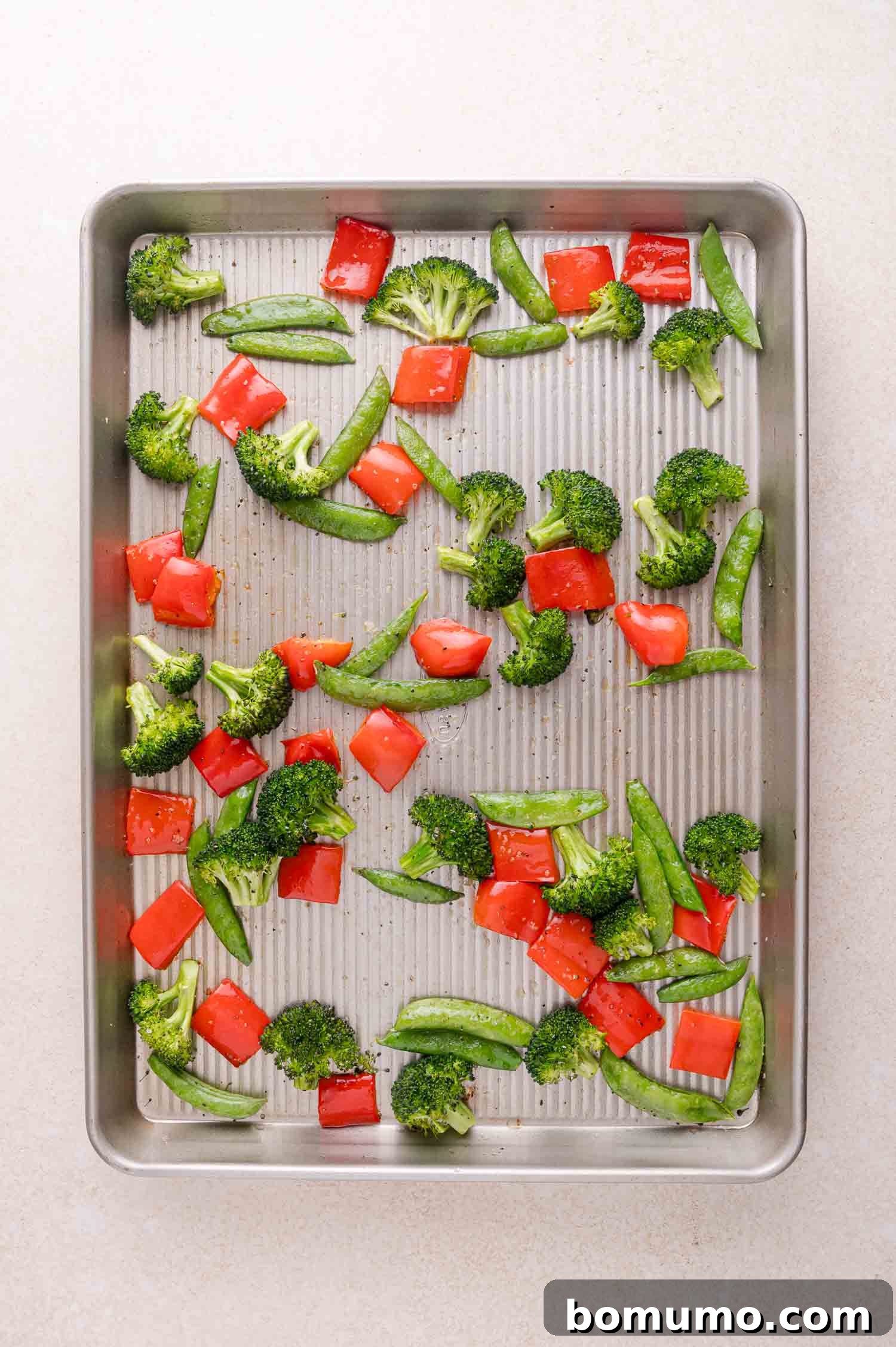 A baking sheet with raw broccoli florets, red bell pepper pieces, and snap peas spread evenly on the surface.