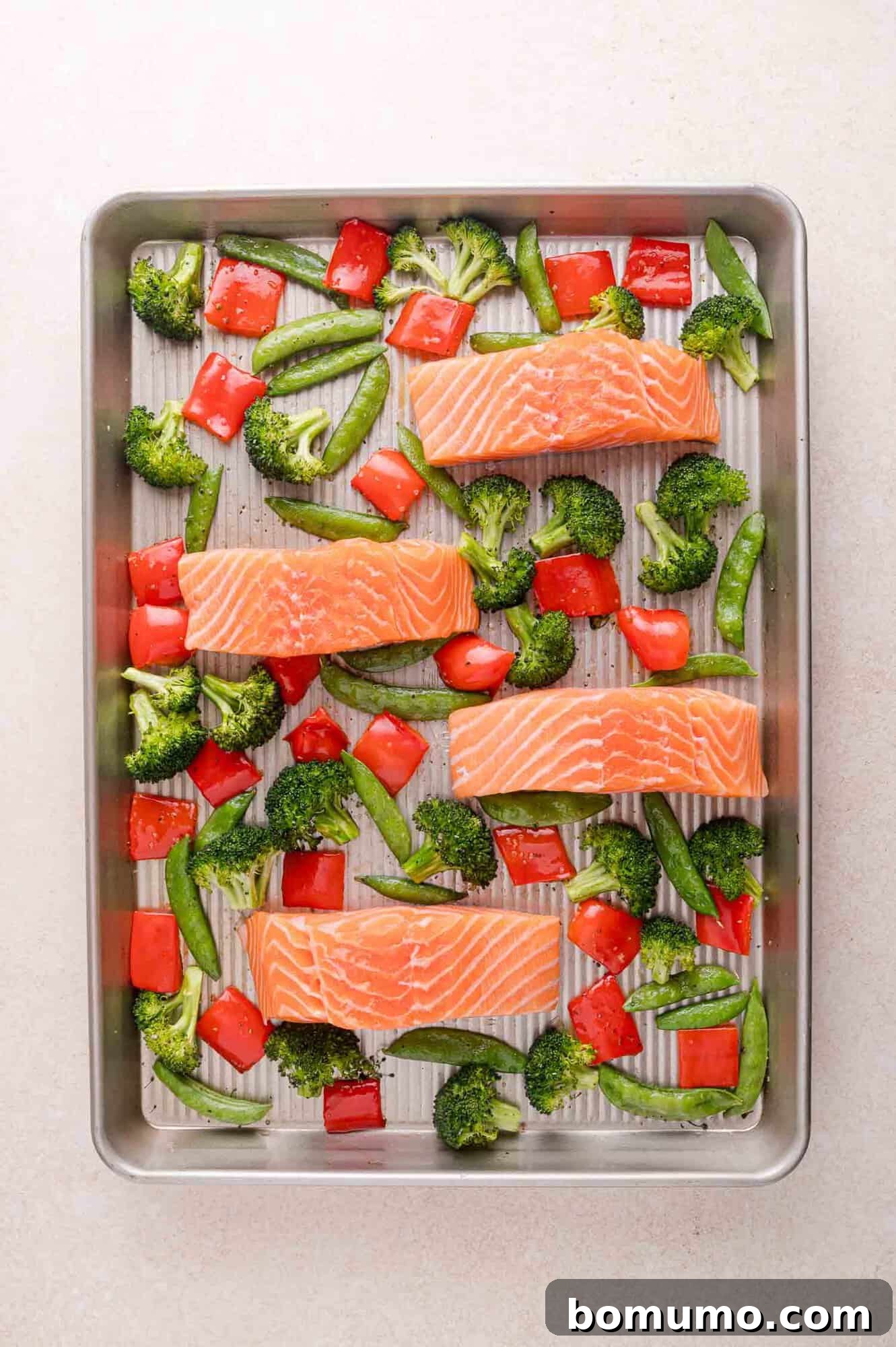Four raw salmon fillets, broccoli, sugar snap peas, and red bell pepper pieces arranged on a metal baking sheet, ready for cooking.