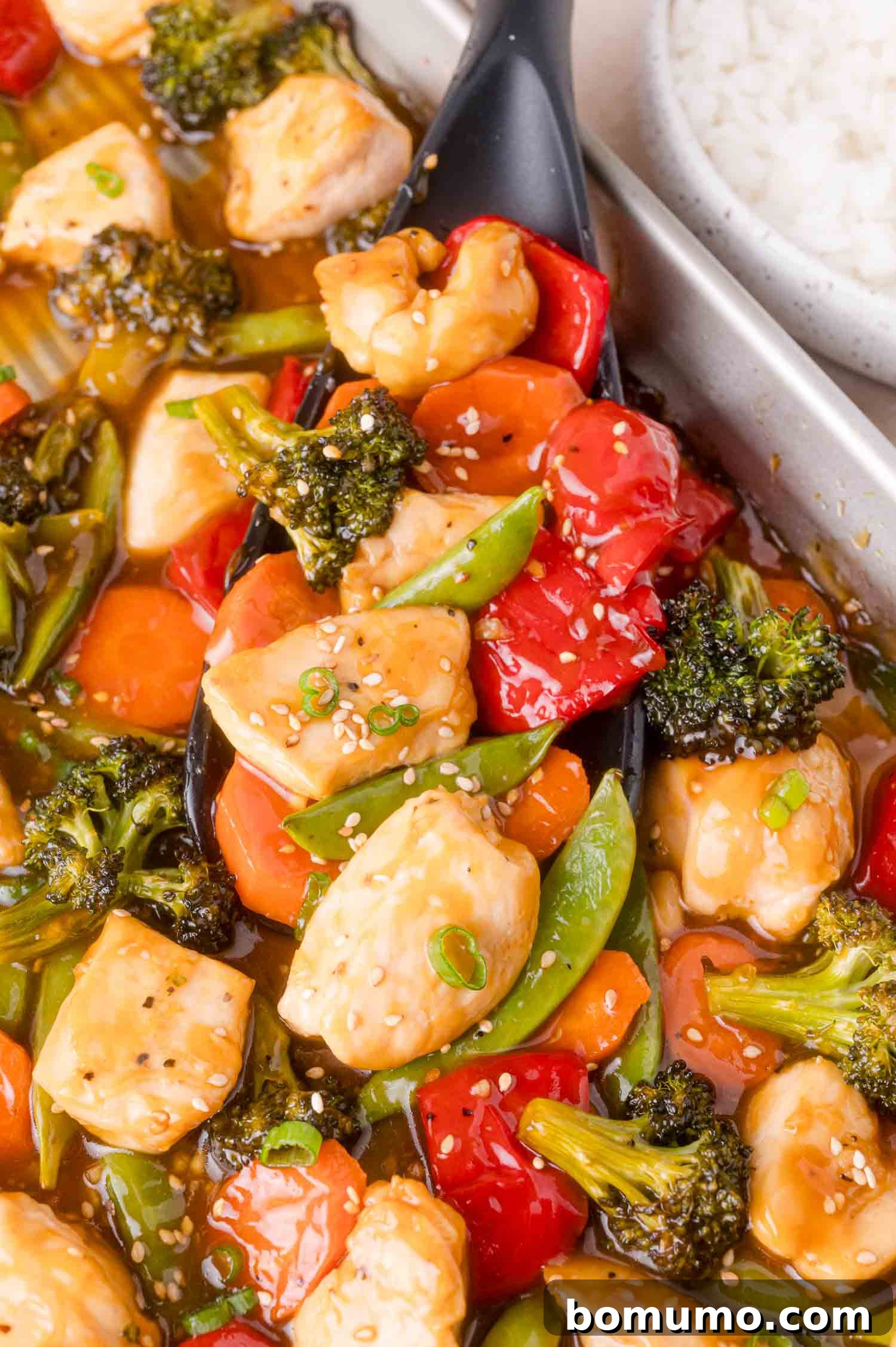 Close-up of a spoonful of chicken stir fry with broccoli, carrots, red bell peppers, and snap peas, topped with sesame seeds, next to a bowl of white rice.