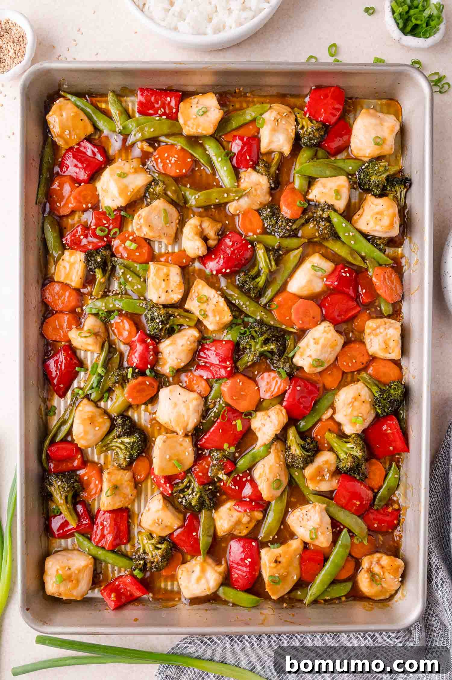 A sheet pan filled with cooked chicken pieces, broccoli, red bell peppers, snap peas, and carrots in a savory sauce. Bowls of rice and green onions are nearby.