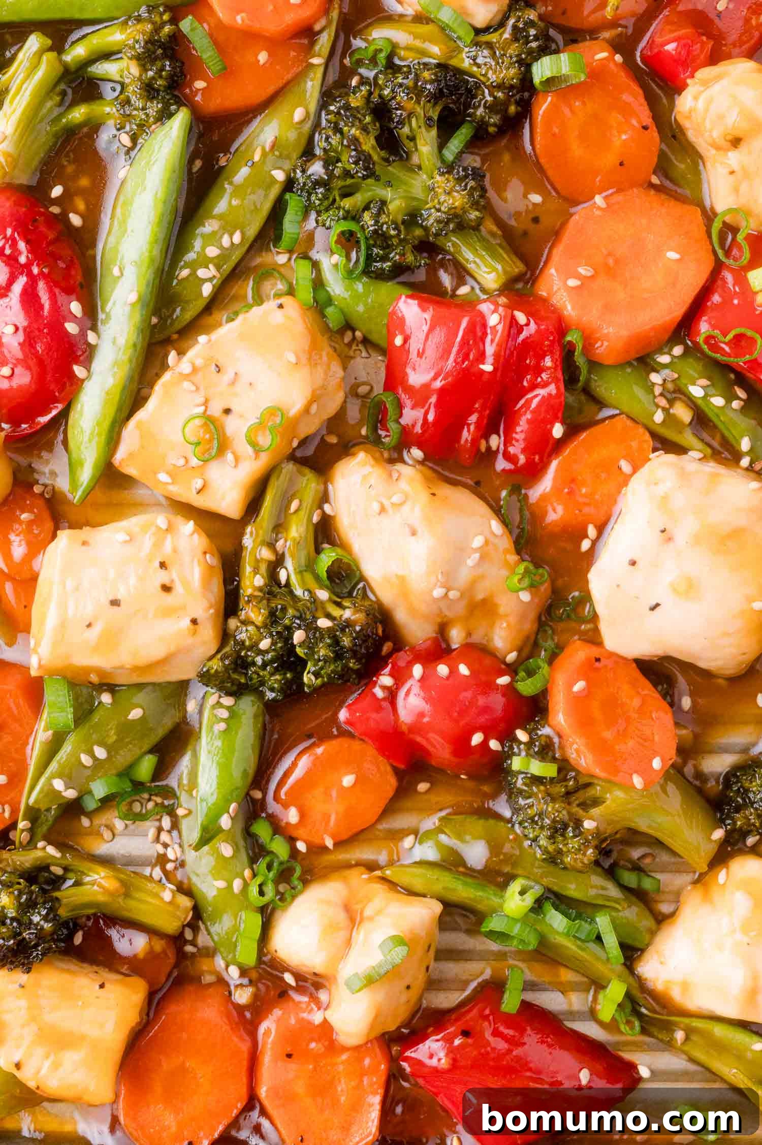 Close-up of a colorful stir-fry with chicken, broccoli, carrots, red bell pepper, snap peas, and cherry tomatoes in sauce, garnished with sesame seeds and chopped green onions.