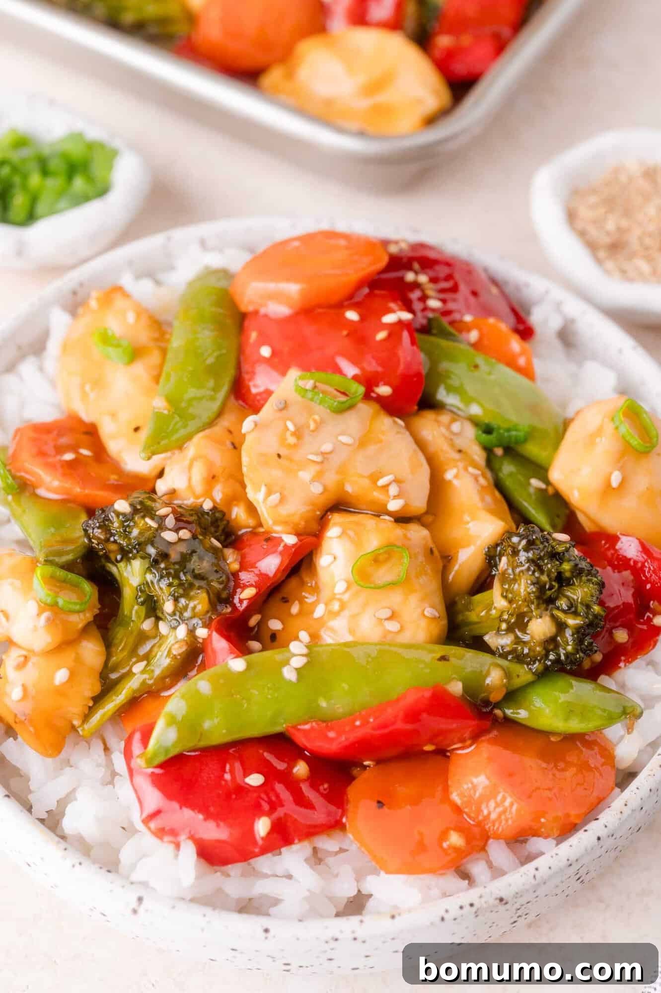 A bowl of white rice topped with stir-fried chicken, broccoli, snap peas, red bell peppers, carrots, and sesame seeds.