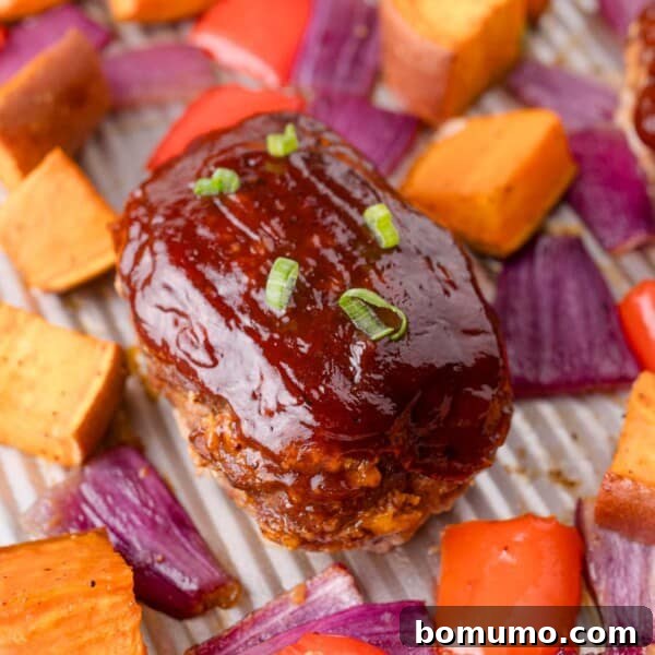 Mini meatloaf topped with barbecue sauce and green onion, surrounded by roasted sweet potatoes, red onions, and red bell peppers on a sheet pan.