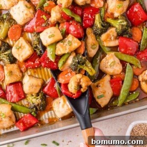 A tray of chicken stir-fry with broccoli, bell peppers, snap peas, carrots, and sauce, being served with a black spatula.