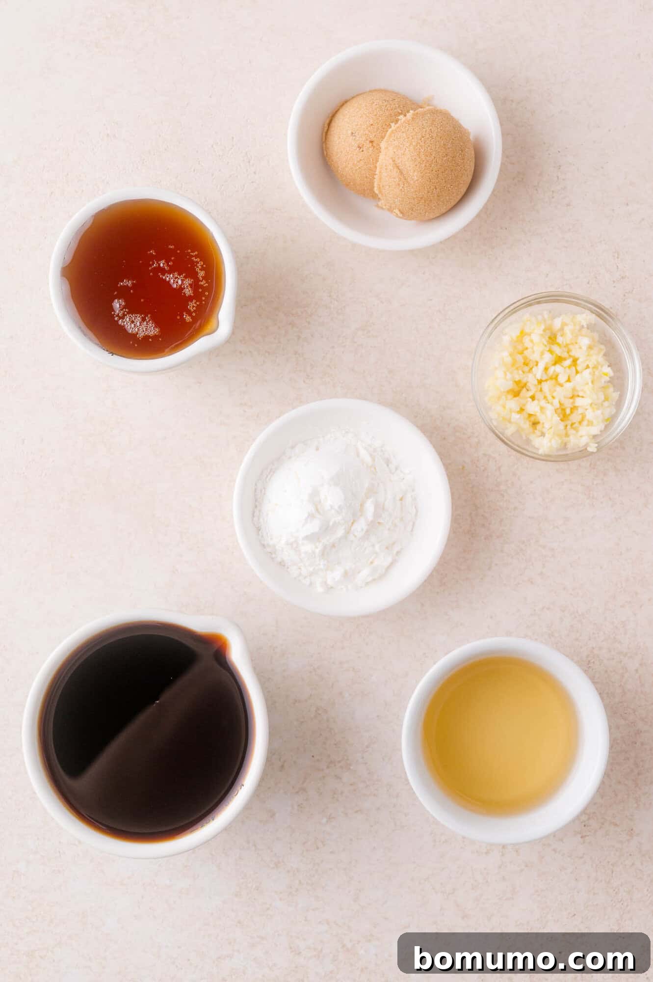 Six small bowls containing soy sauce, brown sugar, minced garlic, honey, cornstarch, and rice vinegar arranged on a light surface.