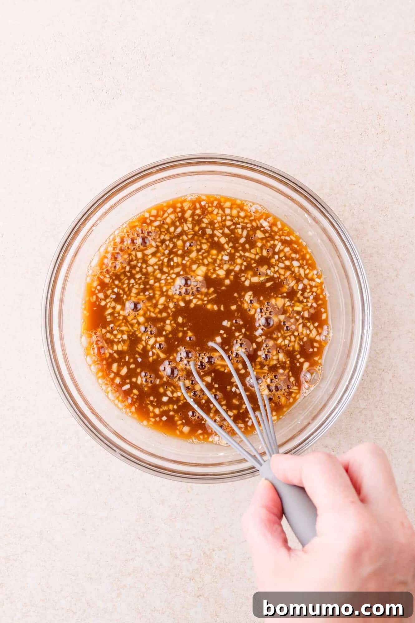 A hand holding a whisk mixes a brown sauce with visible garlic in a clear glass bowl on a light-colored surface.