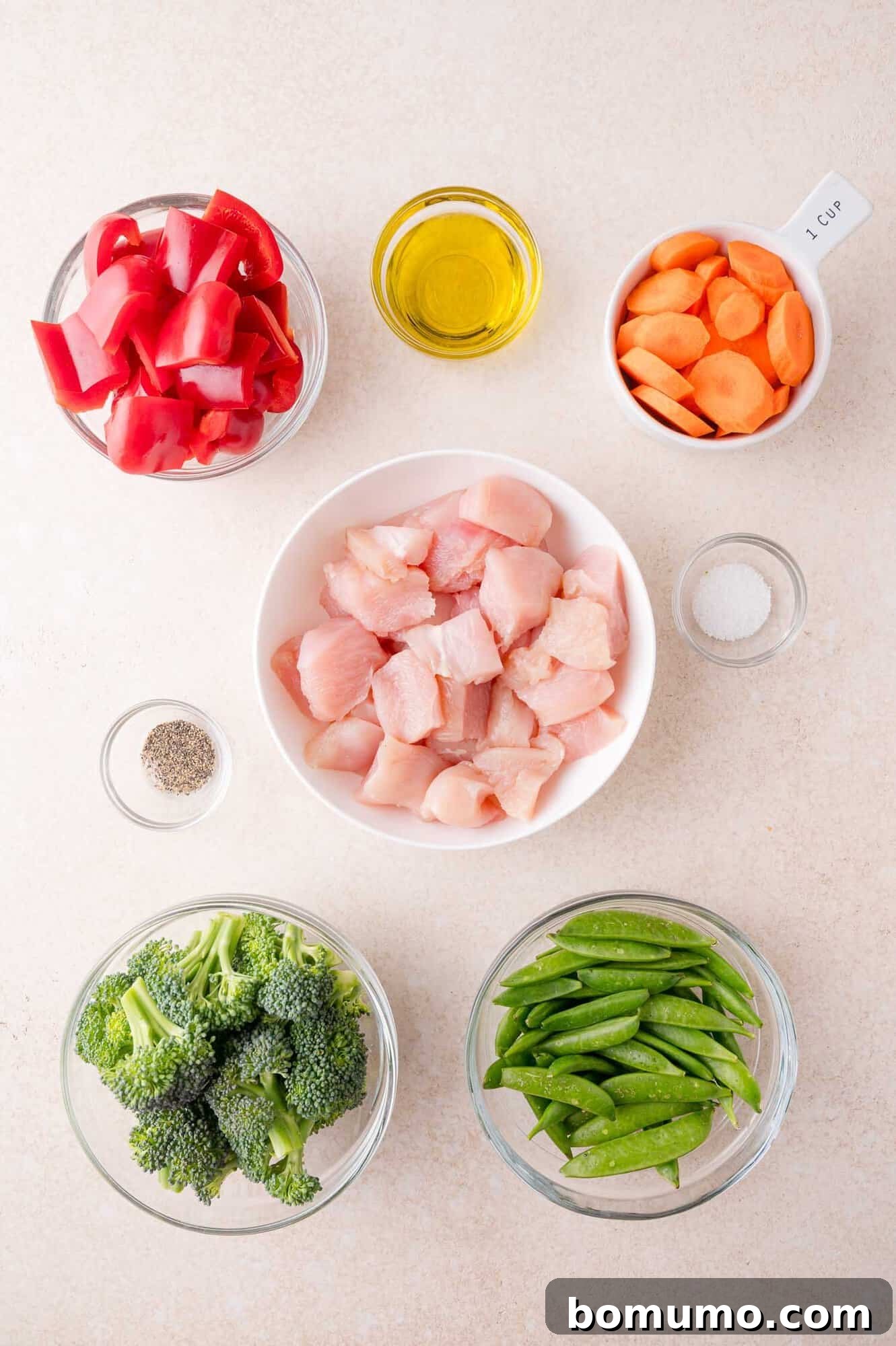 Raw diced chicken, broccoli, snap peas, red bell peppers, sliced carrots, olive oil, salt, and pepper arranged in separate bowls on a light surface.