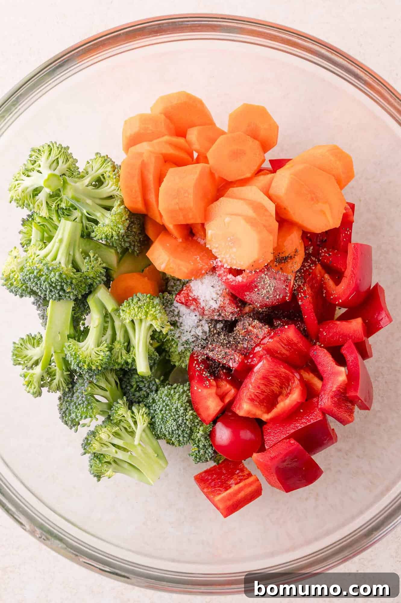 A glass bowl containing chopped broccoli, sliced carrots, and diced red bell pepper, topped with salt and pepper.