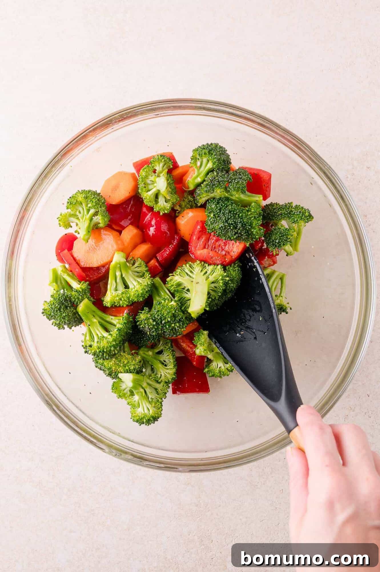 A hand uses a black spatula to mix fresh broccoli, red bell pepper, and sliced carrots in a glass bowl.