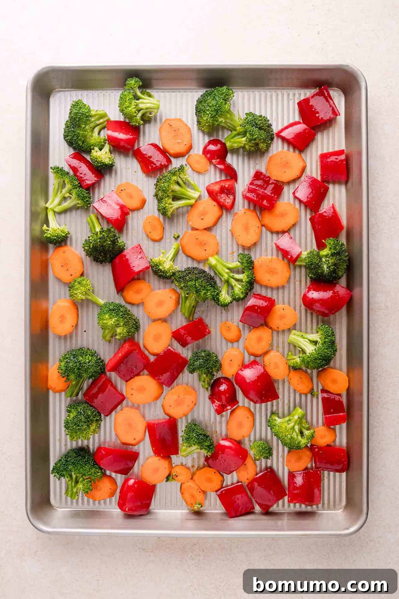 Chopped broccoli, red bell pepper, and carrot pieces are spread evenly on a metal baking sheet, ready for roasting.