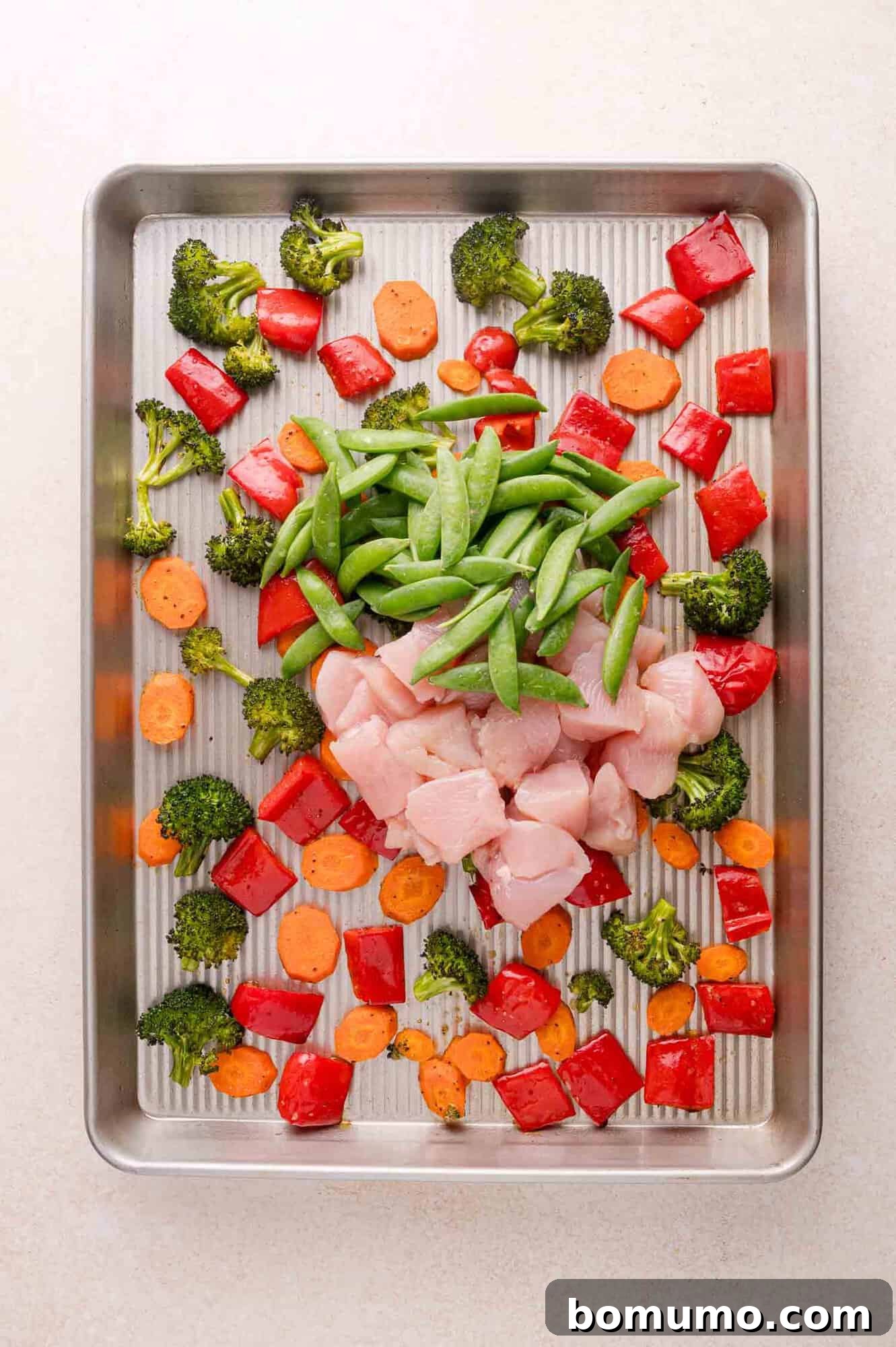 Raw chicken pieces, sugar snap peas, chopped red bell pepper, broccoli florets, and sliced carrots arranged on a metal baking sheet.