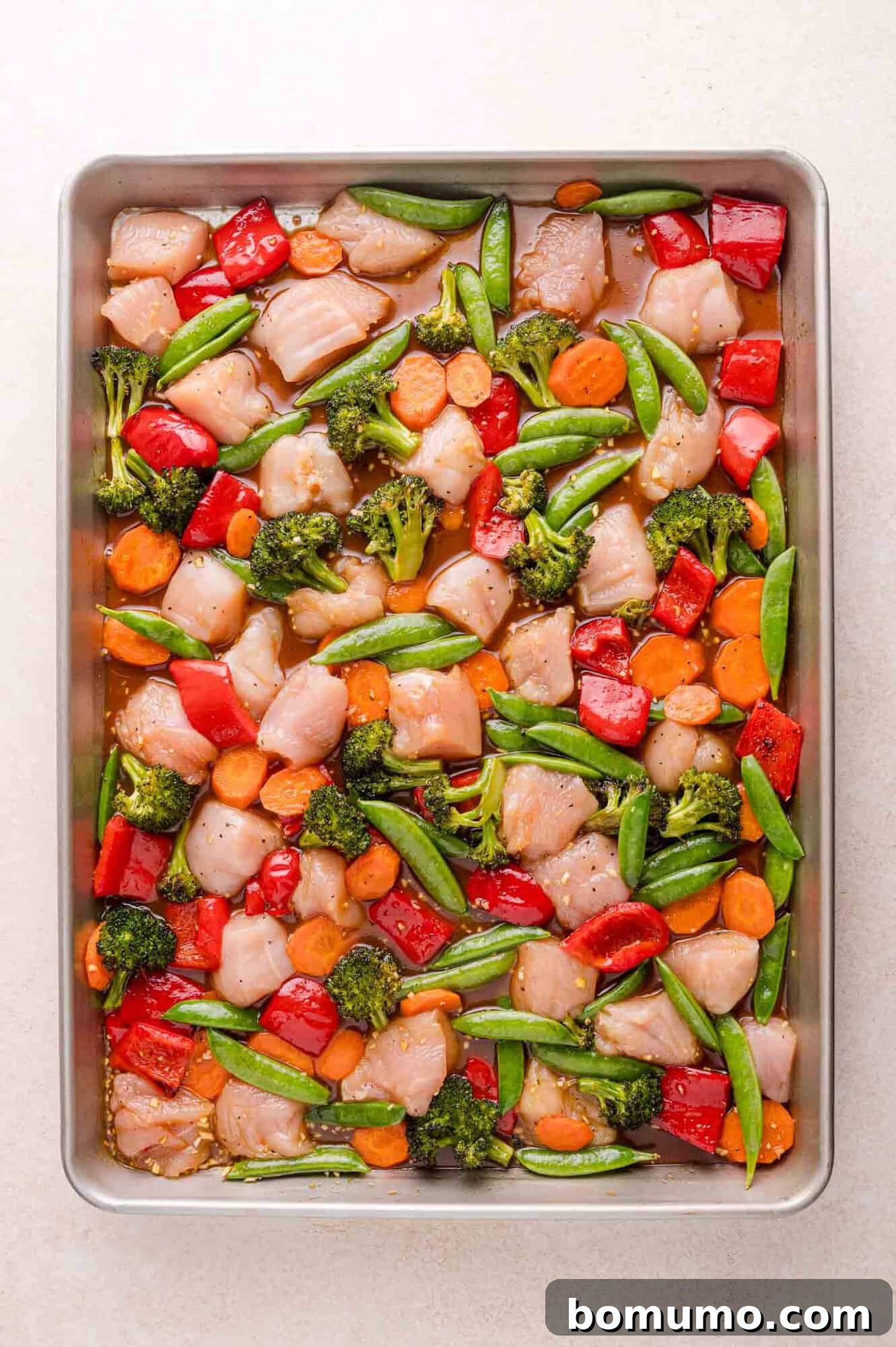 A sheet pan filled with raw diced chicken, broccoli, red bell peppers, snap peas, and carrot slices, ready to be cooked.