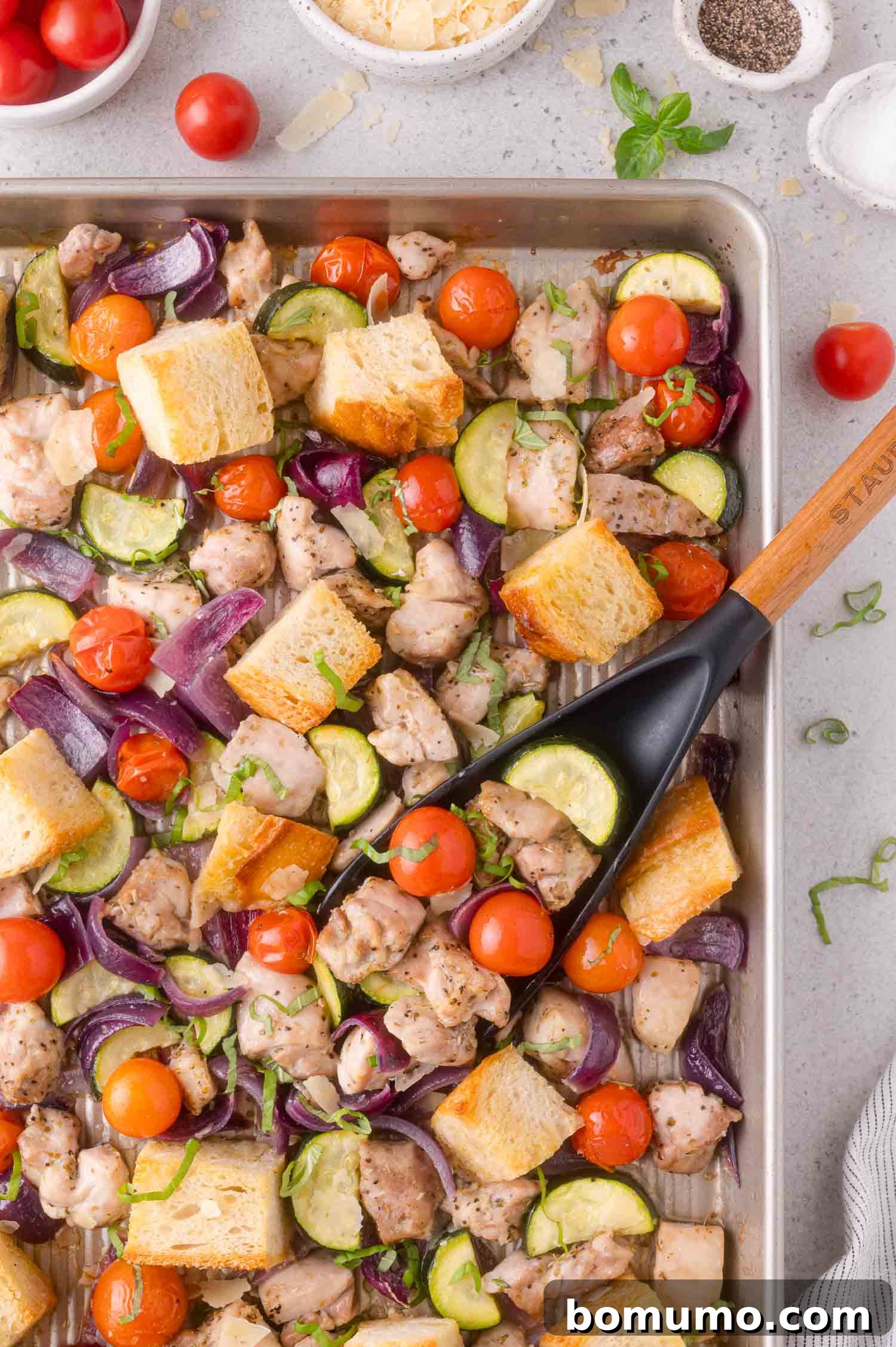 Sheet pan with roasted chicken, cherry tomatoes, zucchini, red onion, and bread cubes, with a spatula lifting a portion.