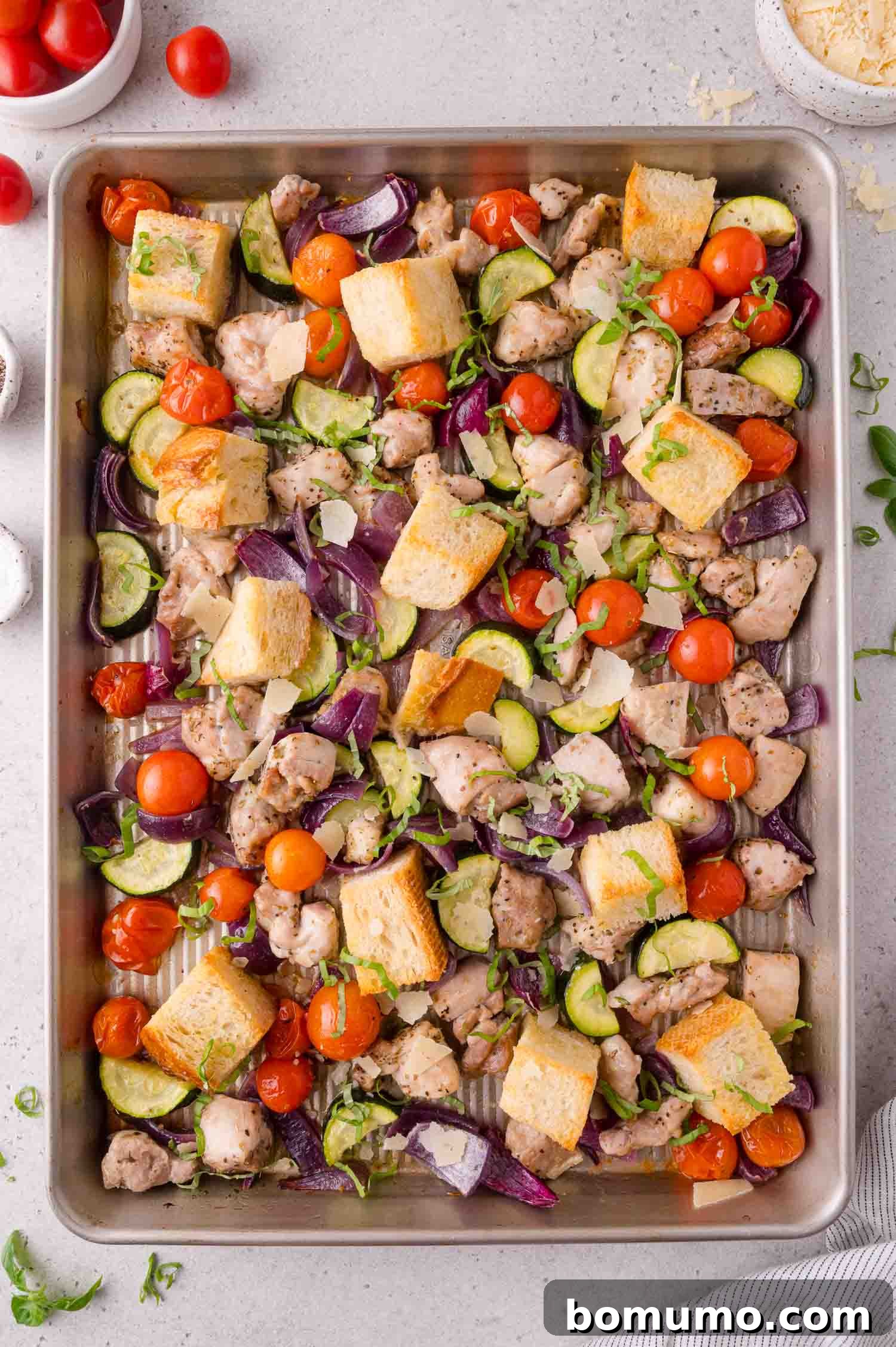 A sheet pan filled with roasted chicken pieces, cherry tomatoes, zucchini, red onion, and cubes of bread, garnished with herbs.