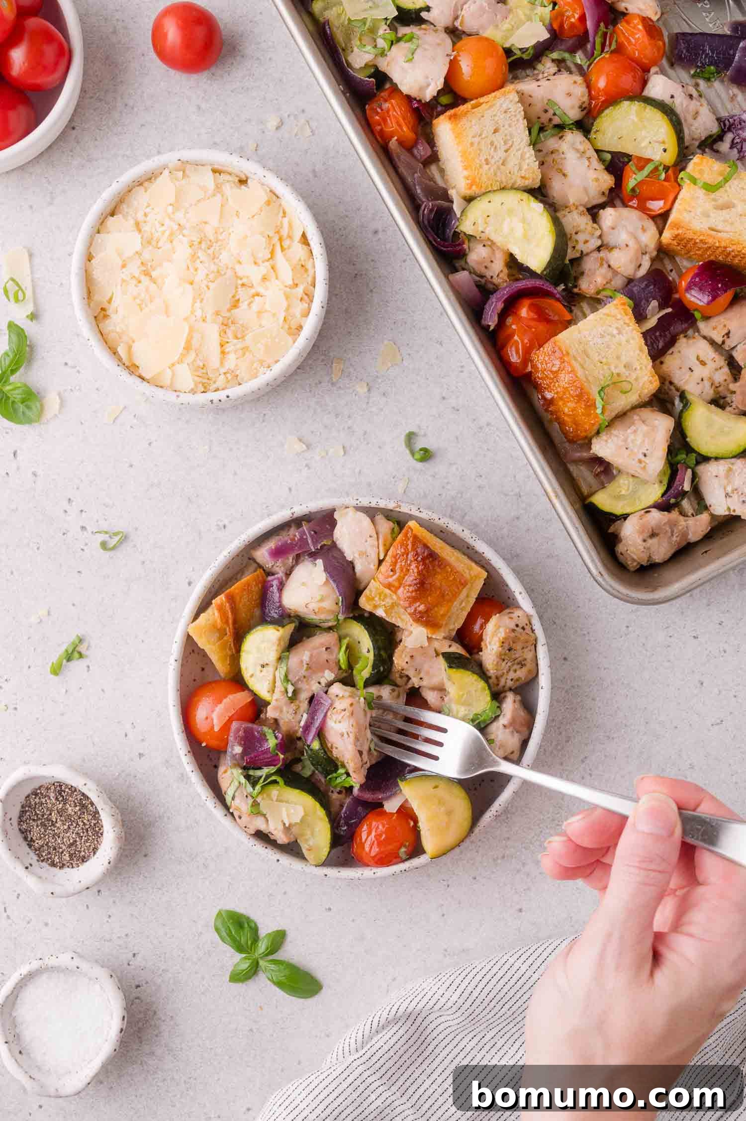 A hand holds a fork over a bowl of salad with chicken, bread cubes, zucchini, tomatoes, and red onions. Nearby are bowls of shredded cheese, salt, pepper, and a tray of the same salad.
