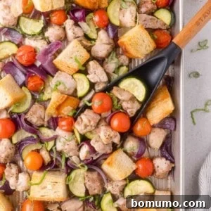 A sheet pan with baked chicken pieces, zucchini, cherry tomatoes, red onion, and bread cubes, garnished with fresh herbs and a black serving spatula.