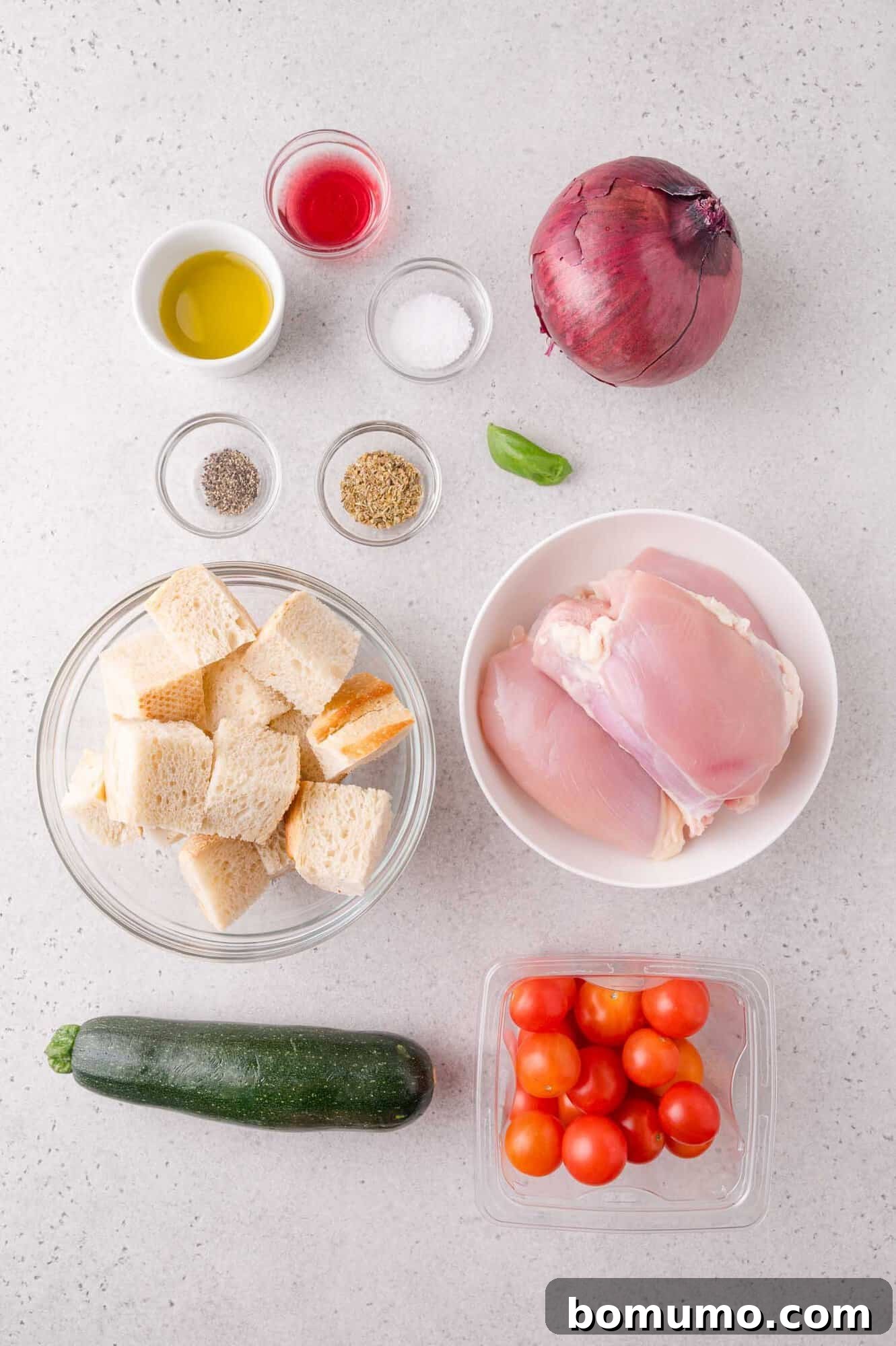 Ingredients on a countertop, including chicken thighs, cubed bread, cherry tomatoes, zucchini, red onion, basil, olive oil, vinegar, salt, pepper, and dried herbs.